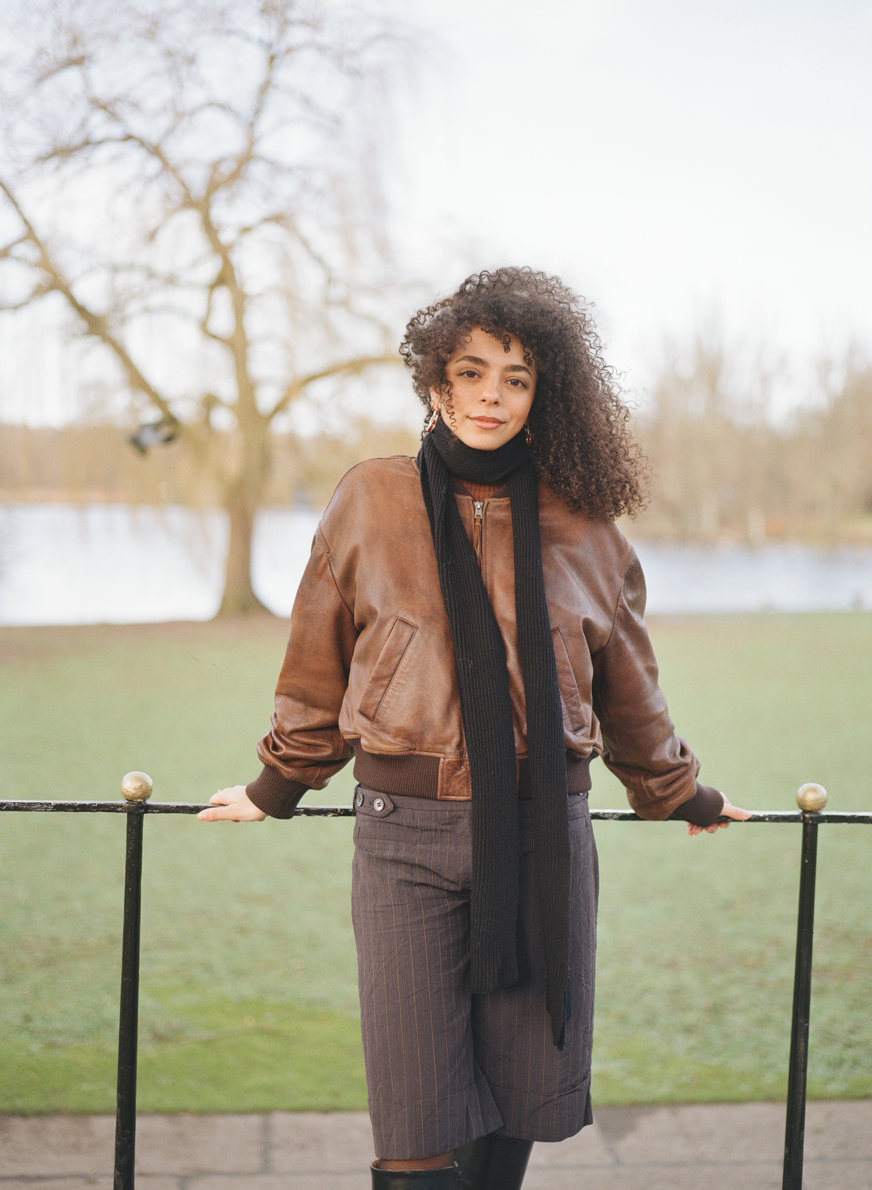 A person with curly hair wearing a brown jacket and black scarf, standing on a bridge overlooking a grassy field.