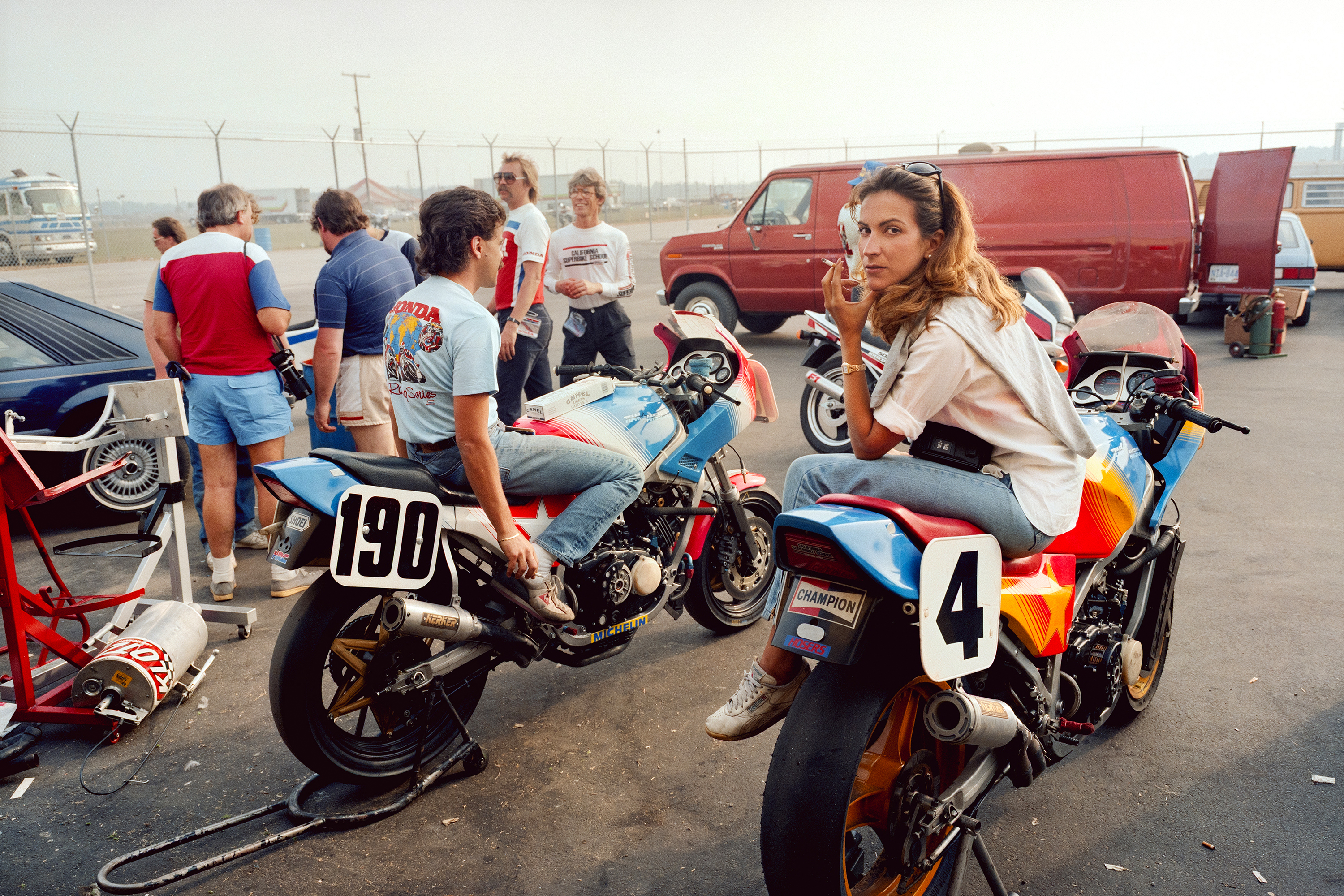 Woman on racing motorcycle with number 4, pit area with mechanics working on bike numbered 190, vintage racing scene.