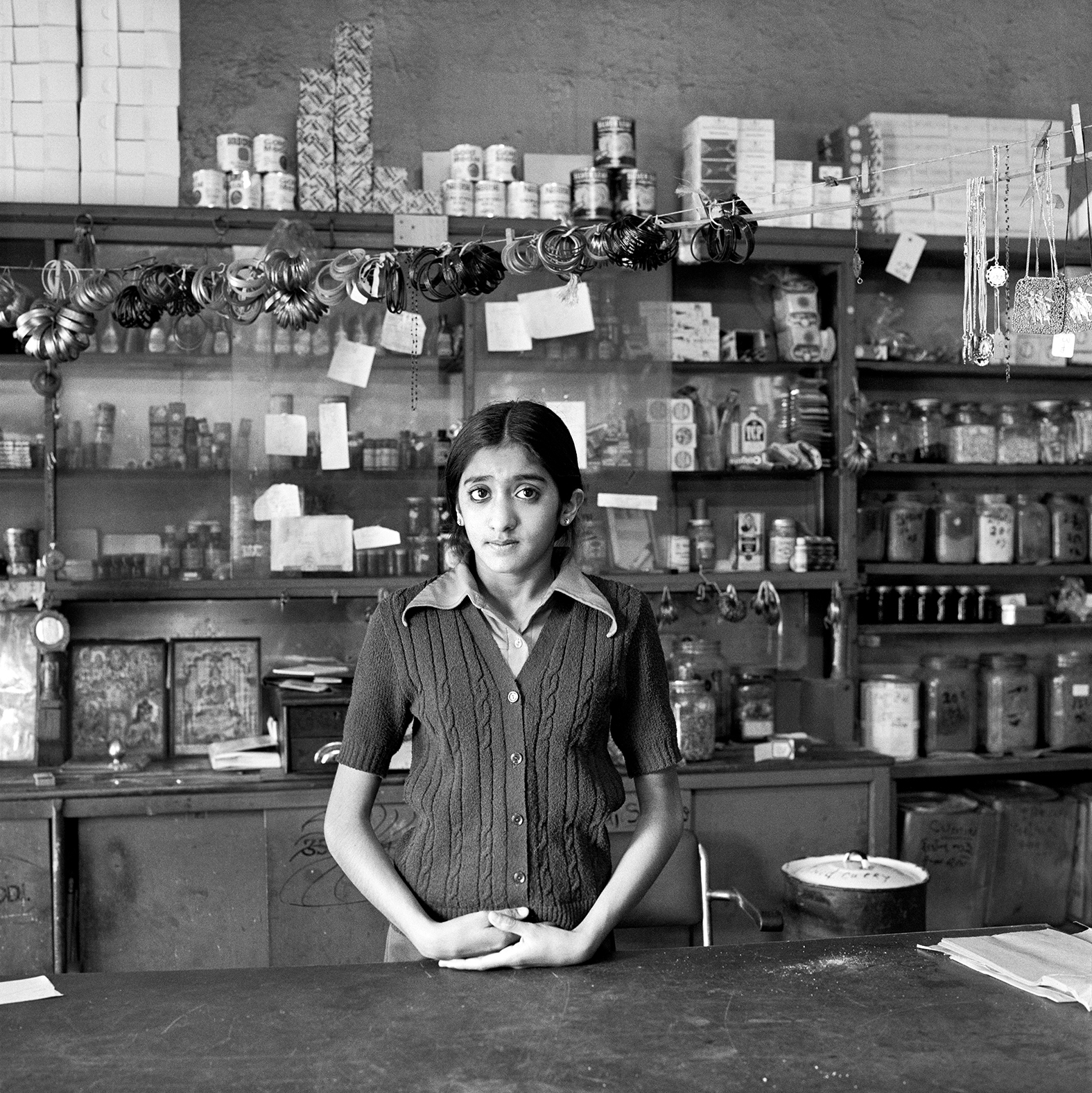 Black and white image of woman standing behind counter in shop with shelves of bottles, jars, and products arranged on wall behind her.