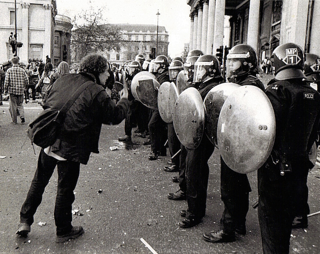 Crowd of protesters facing riot police with shields in a city street, black and white image