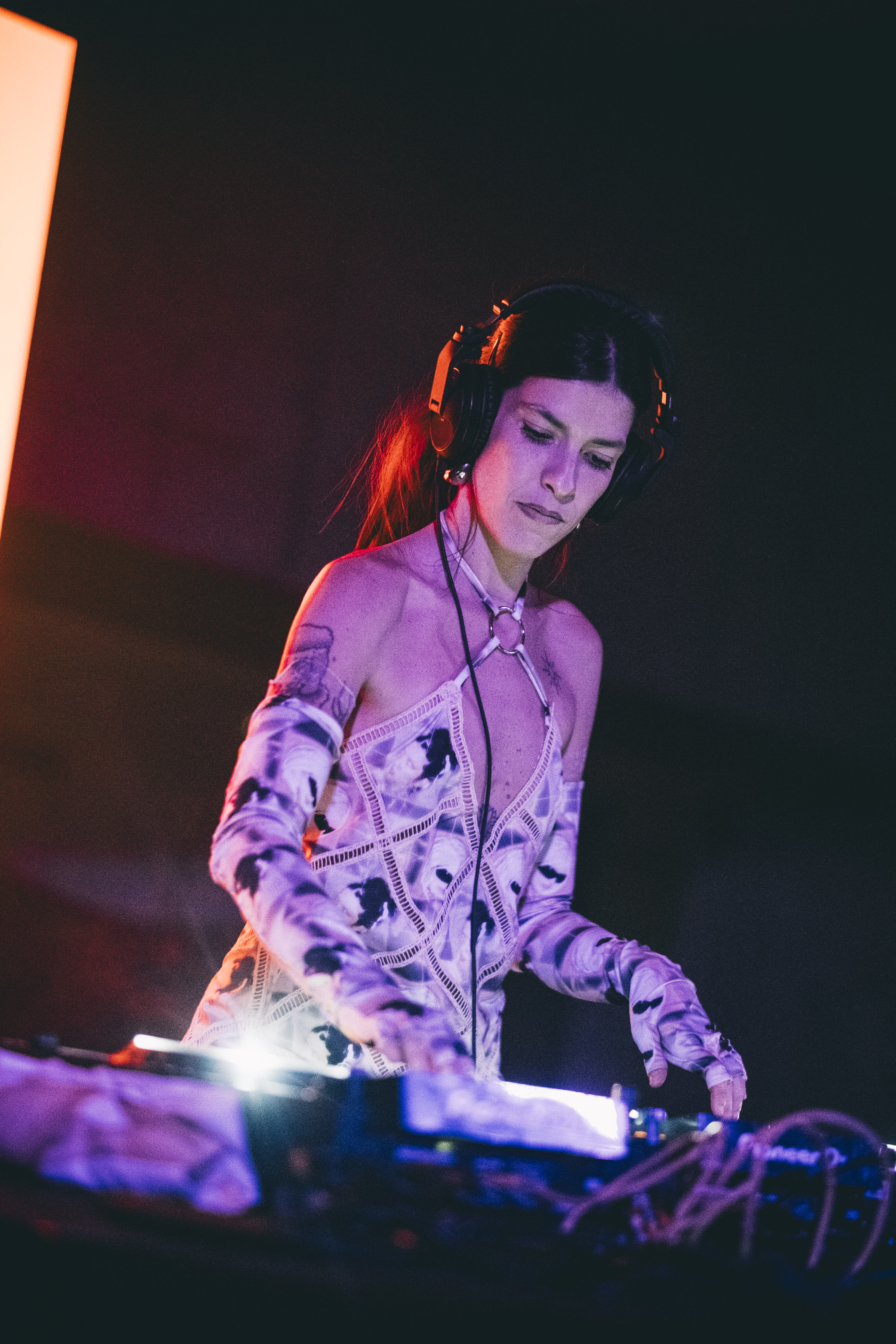 Person in pink top, headphones, and colourful patterned jacket operating sound equipment on a dark stage.