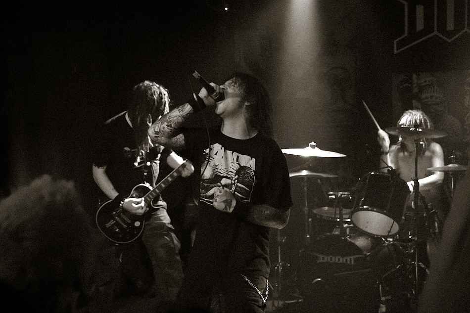 Three musicians performing on stage in dramatic lighting - guitarist on left, vocalist at centre microphone, drummer on right with cymbals visible.