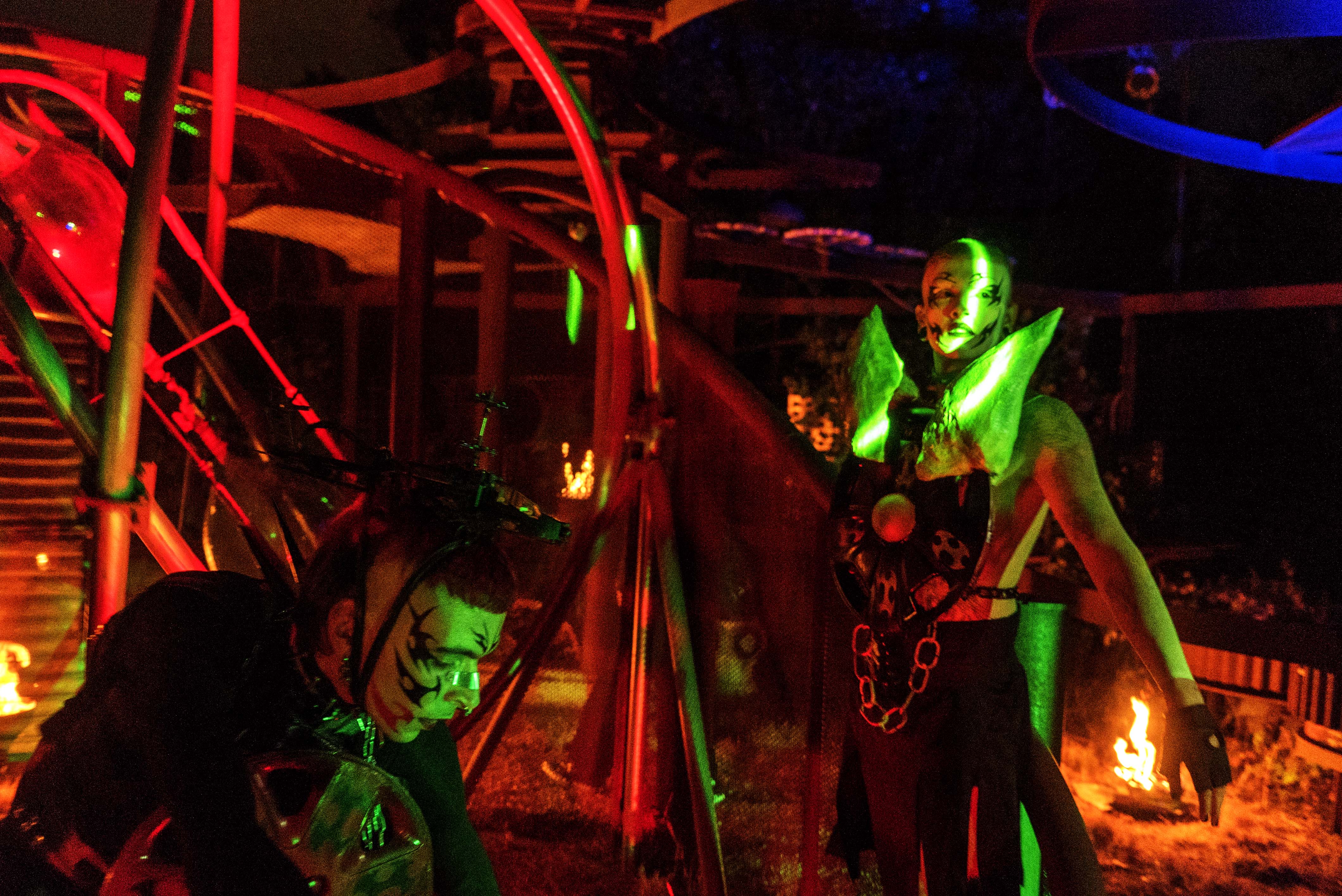 Two people in costumes illuminated by red and green lighting in dark indoor space with metal framework structures.