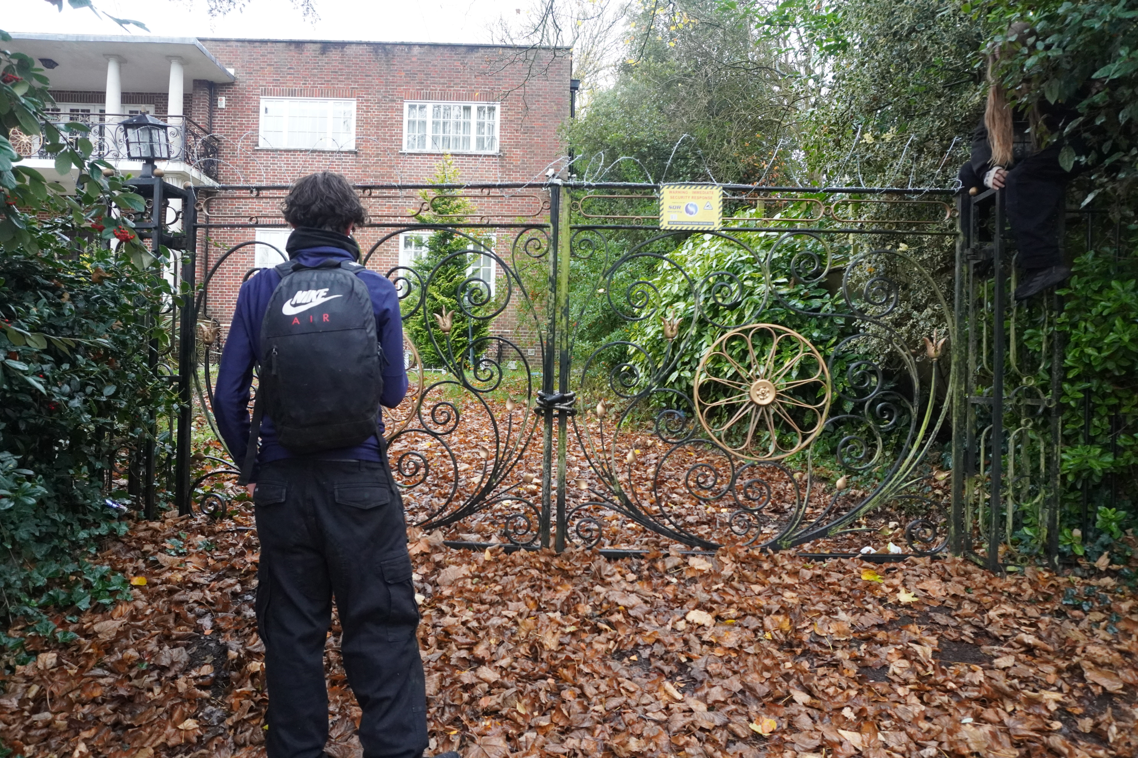 Person in blue overalls cleaning ornate metal gate with decorative spiral patterns, autumn leaves scattered on ground, brick house visible behind.