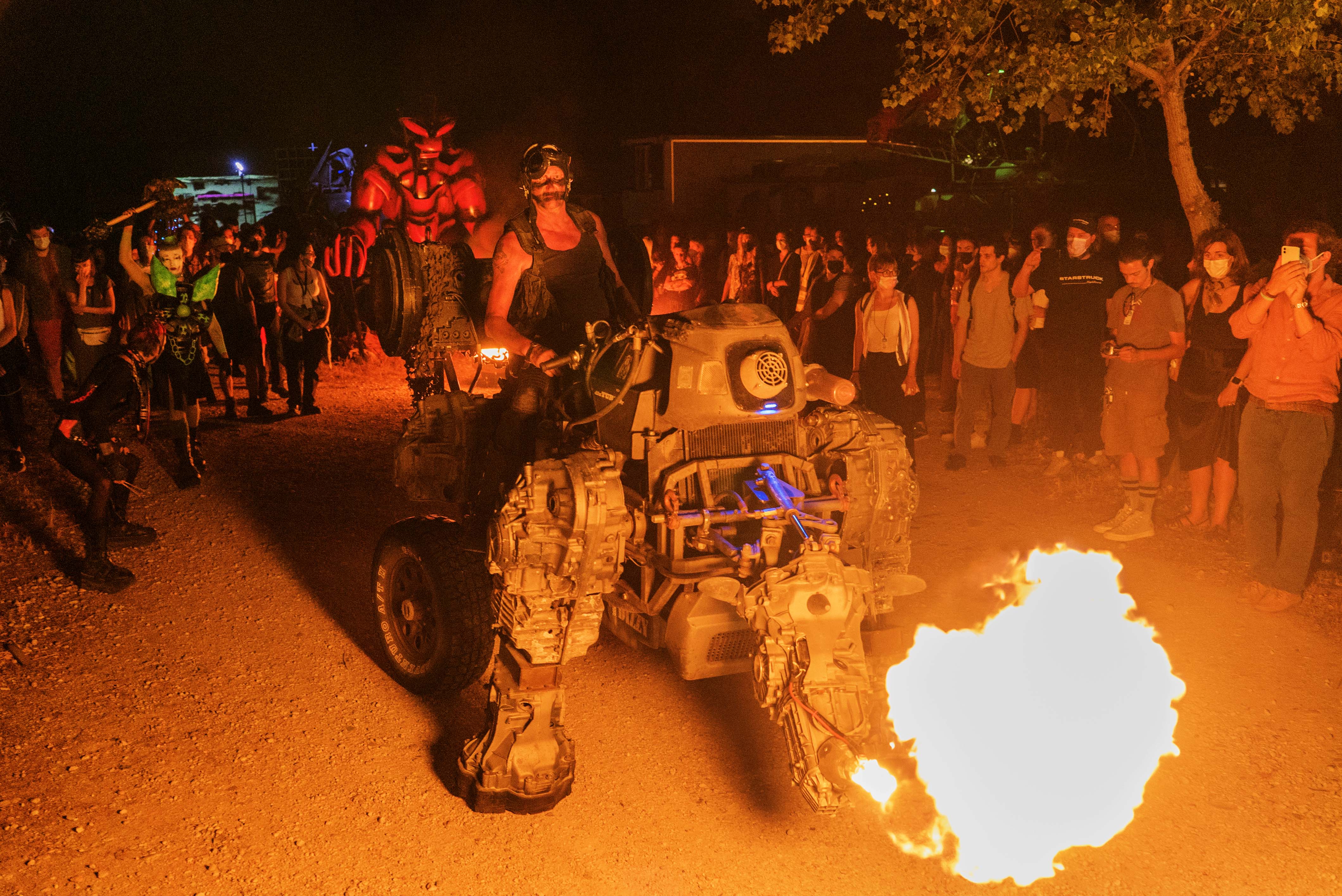 Crowd gathered round motorcycle stunt with bright orange flames and smoke at night-time outdoor event.