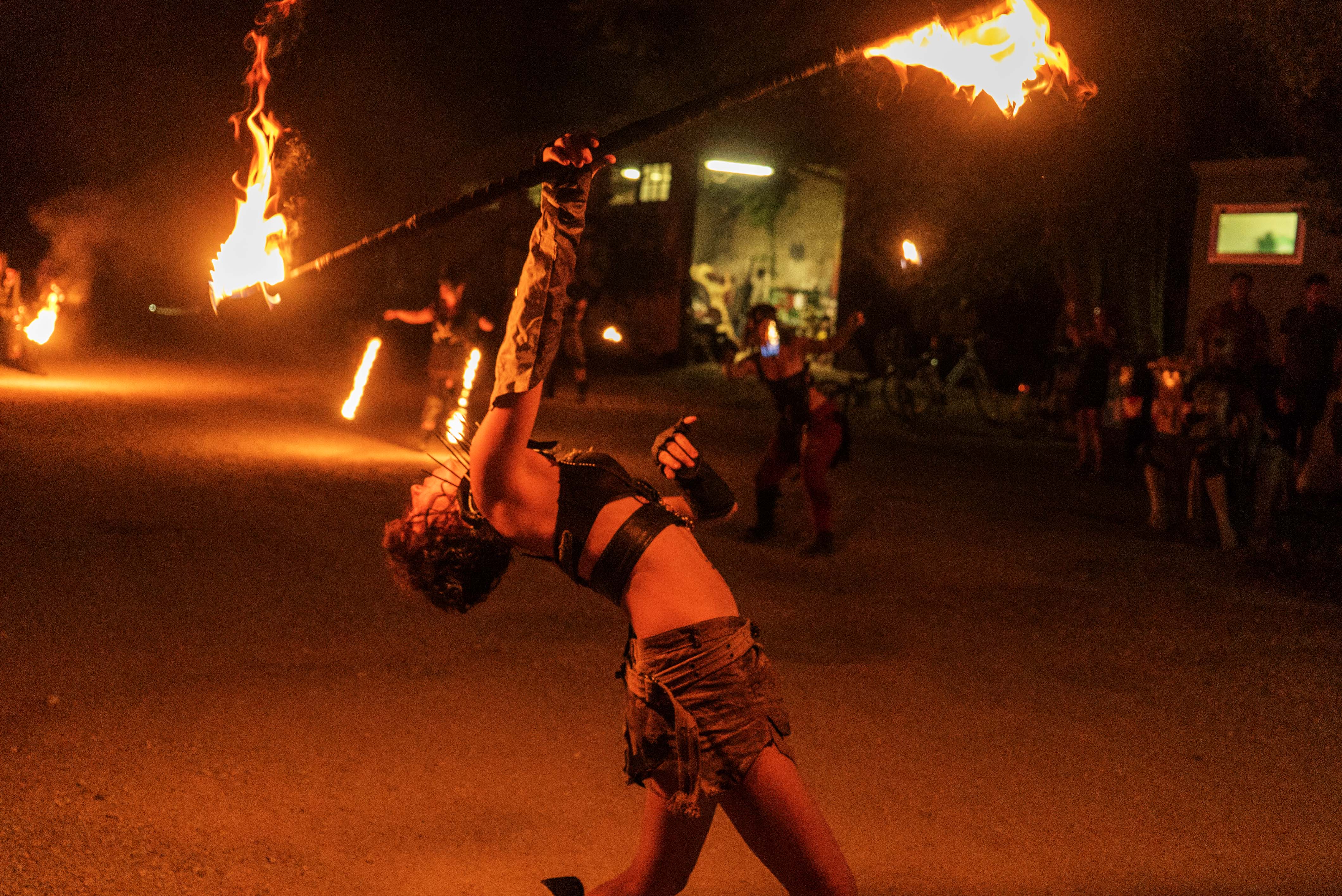 Fire performer swinging flaming torch at night event, crowd watching in background, orange flames against dark sky.