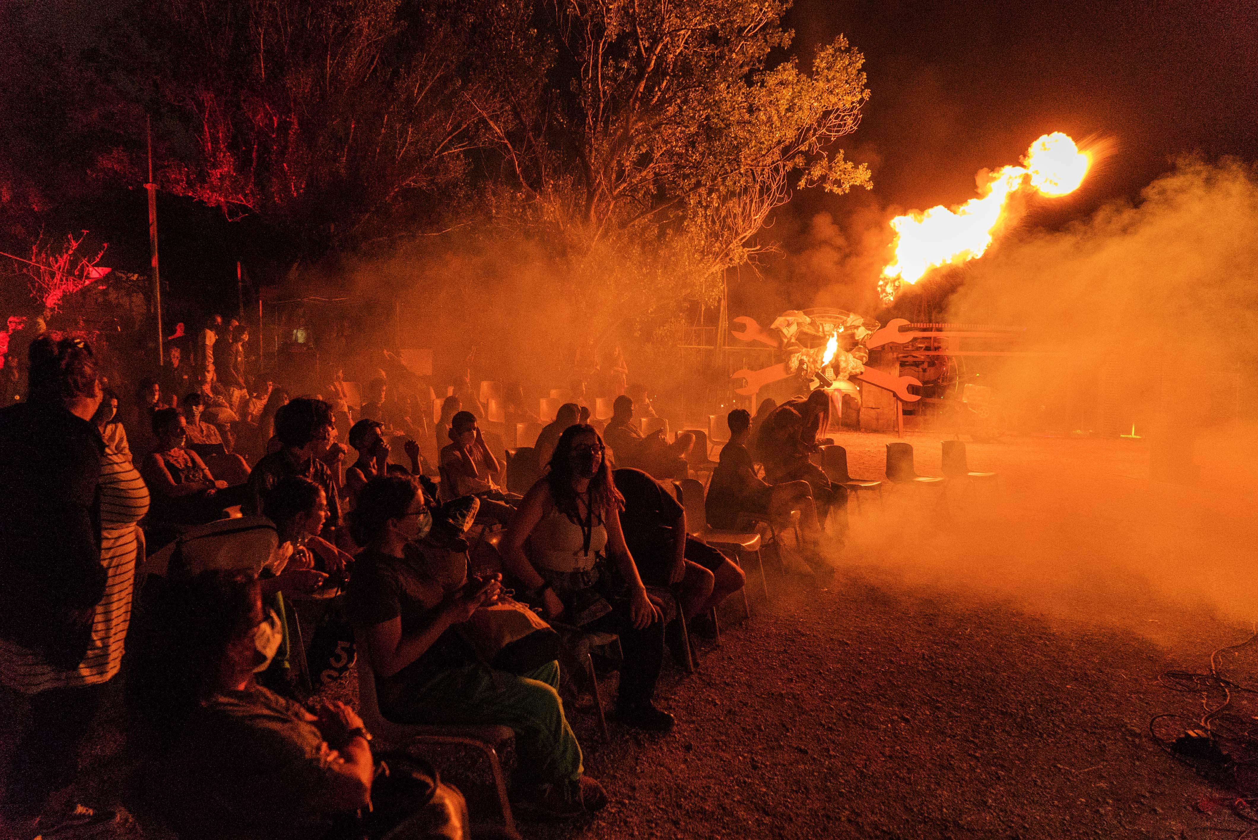 Crowd of people silhouetted against bright orange flames and smoke on a street at night with trees and buildings visible.