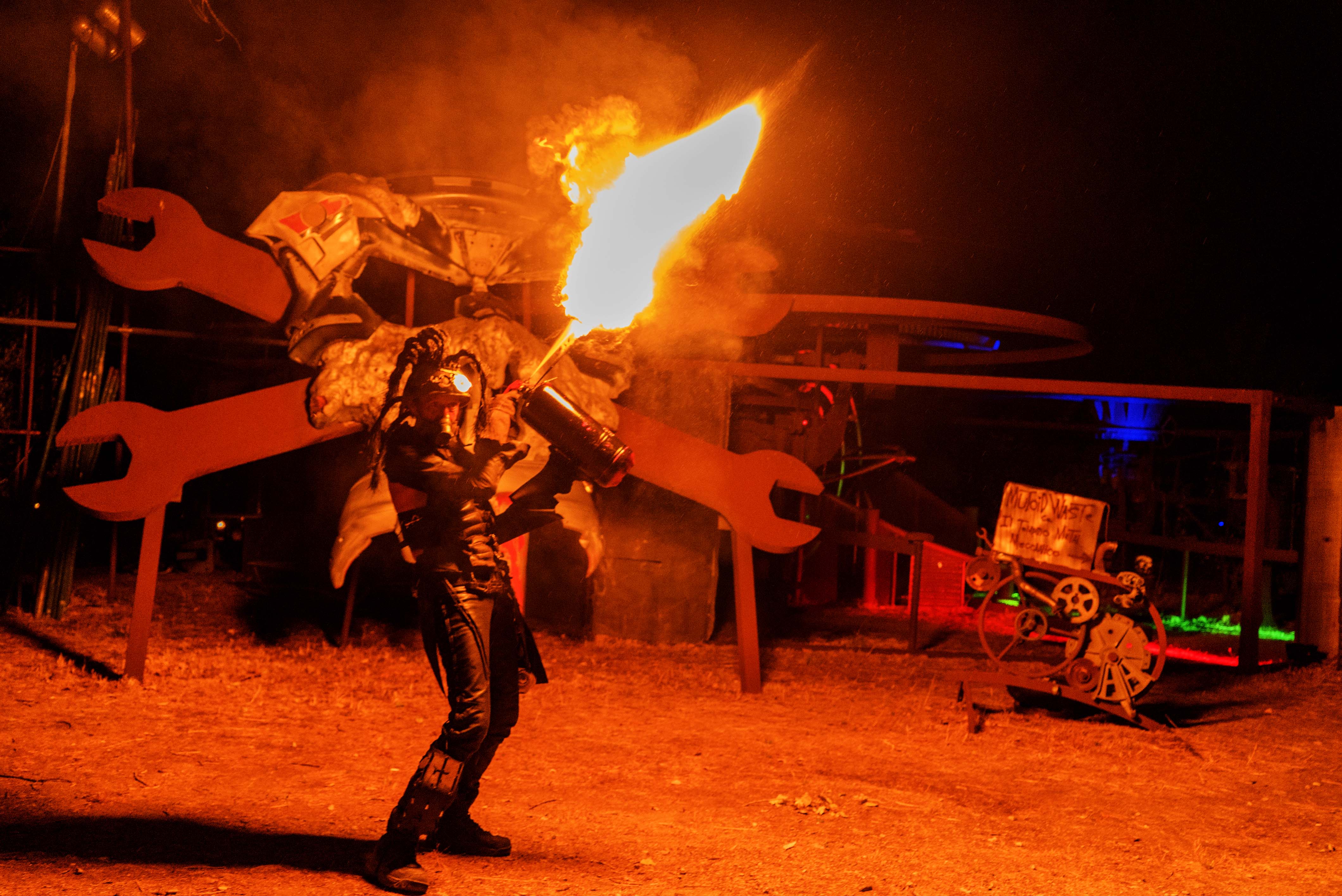 Fire performer in metallic costume spinning large flame torch on industrial stage with orange and red lighting effects.