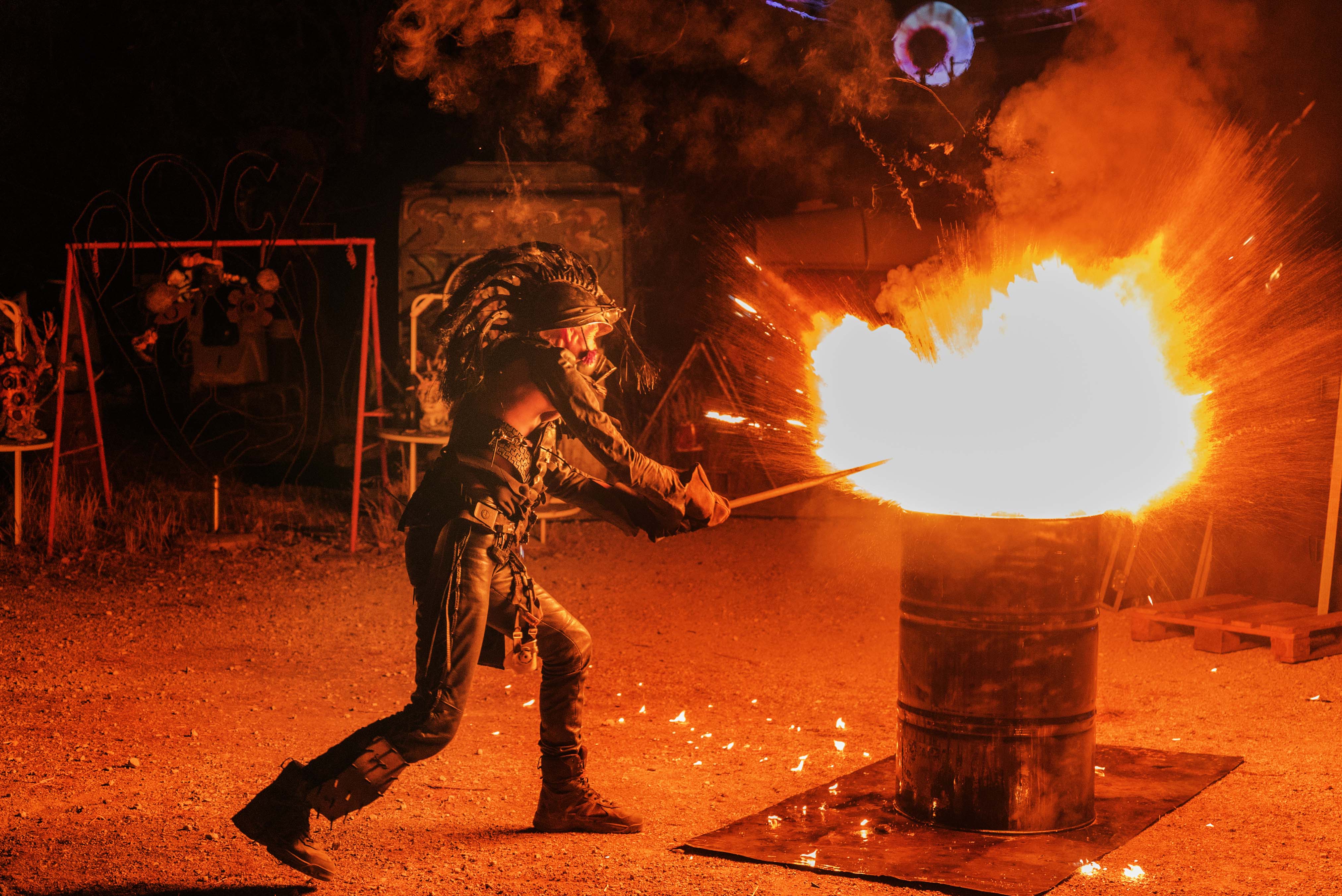 Firefighter in protective gear using water hose on large barrel fire with bright orange and white flames at night training facility.