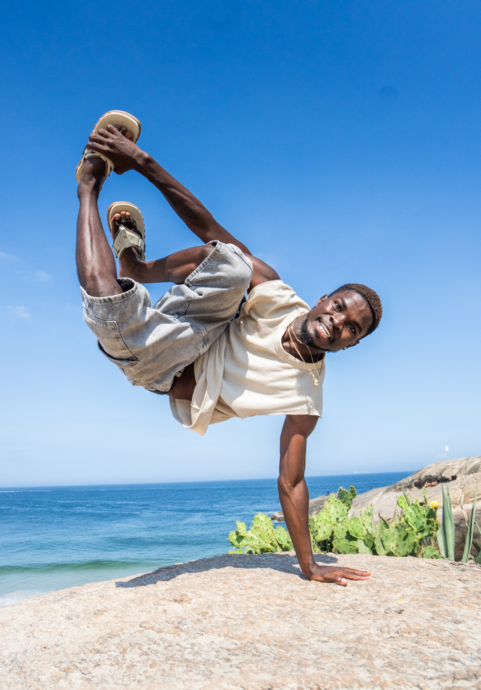 Man performing high kick martial arts move on beach, wearing white gi, blue sky and ocean in background.