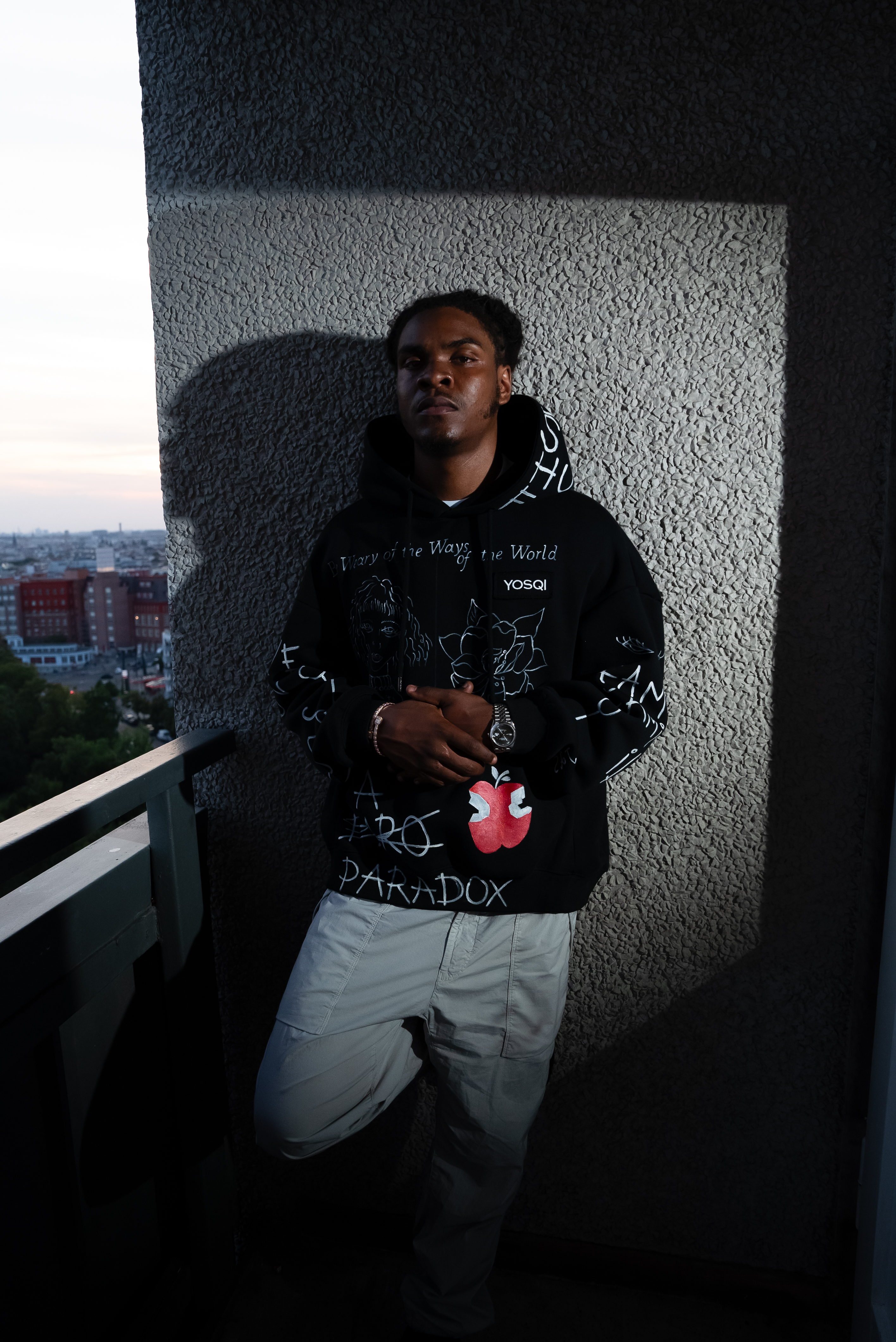 Man in black hoodie and light blue jeans standing on balcony against textured white wall, city view visible in background.