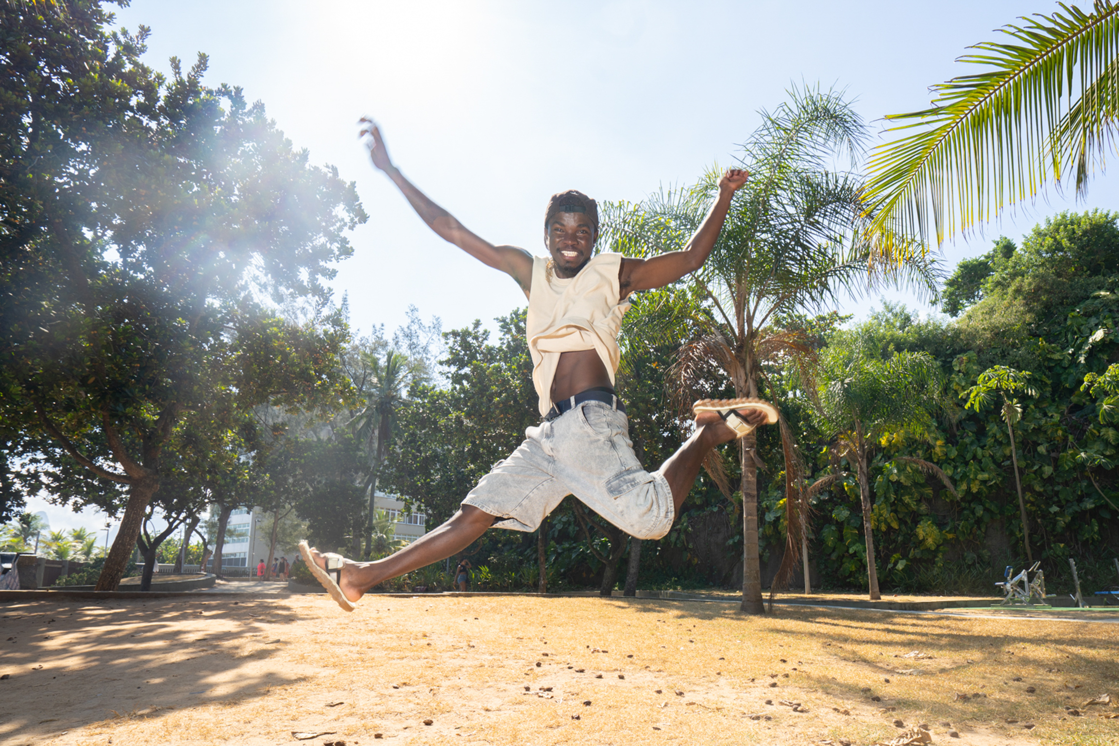 Young boy mid-jump with arms raised, wearing yellow shirt and white shorts, on sandy ground with palm trees and tropical vegetation.
