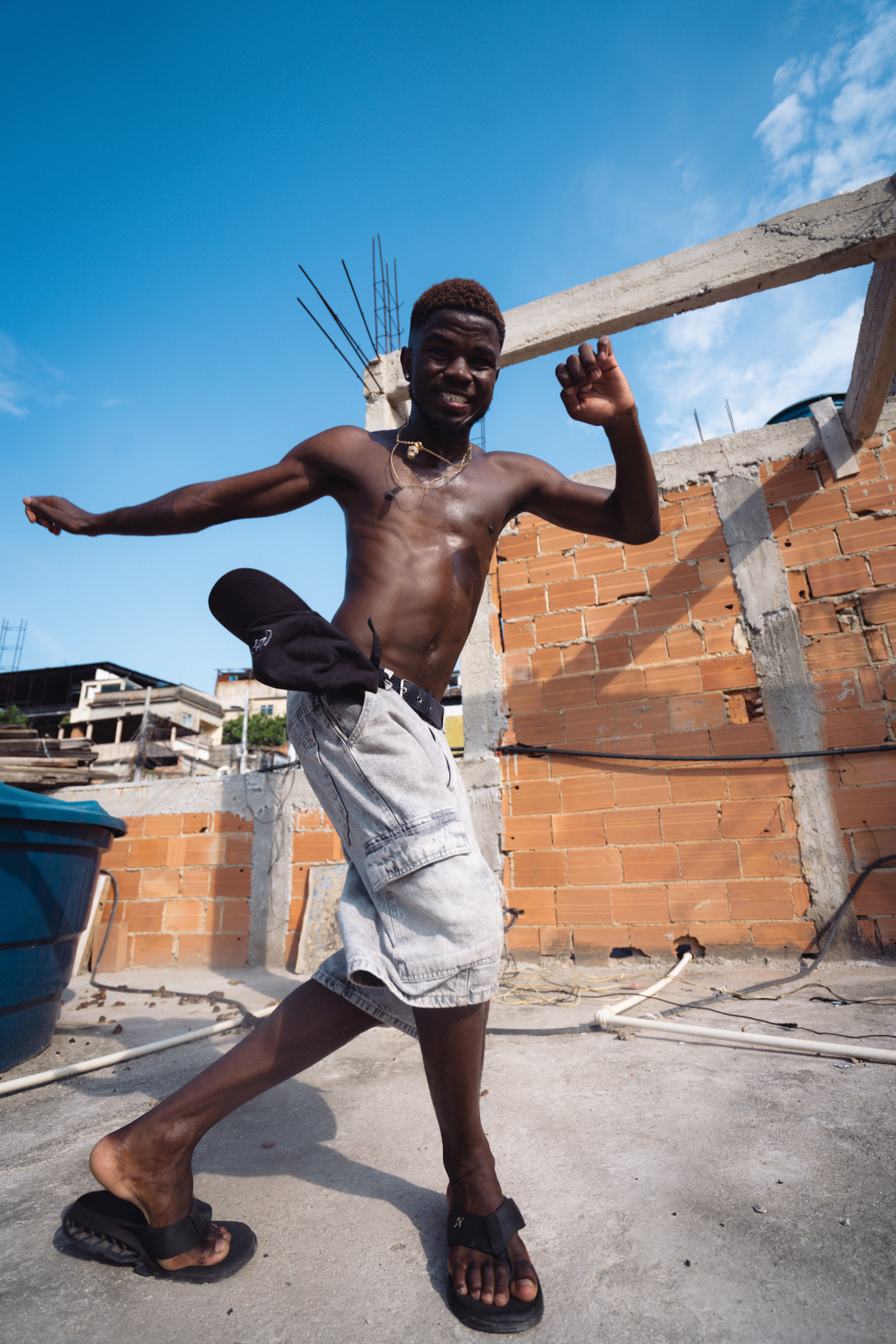 Shirtless young man in grey shorts and sandals jumping with arms outstretched on construction site with brick buildings and blue sky.