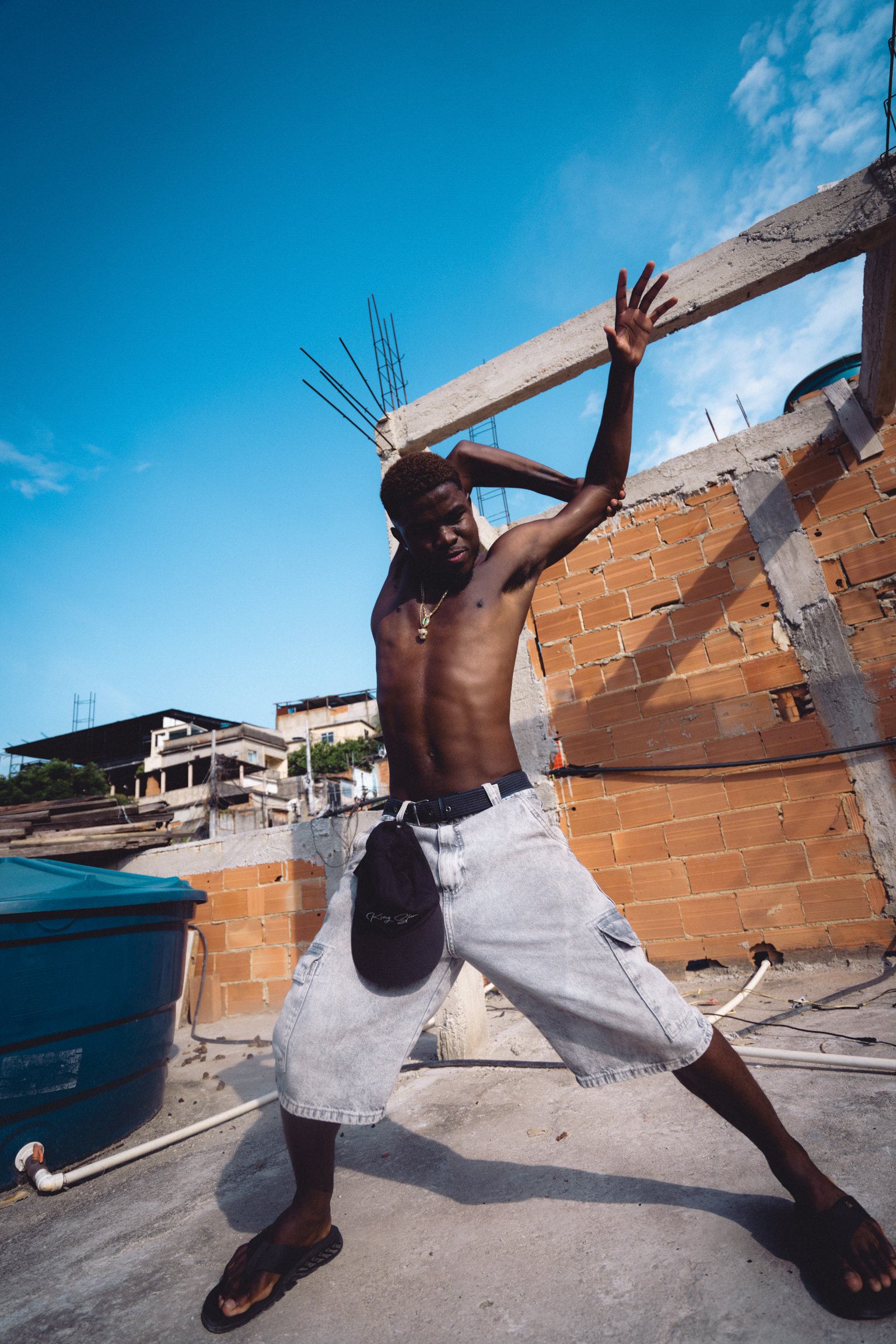 Shirtless man carrying concrete beam on construction site with brick walls, blue sky and unfinished buildings in background.