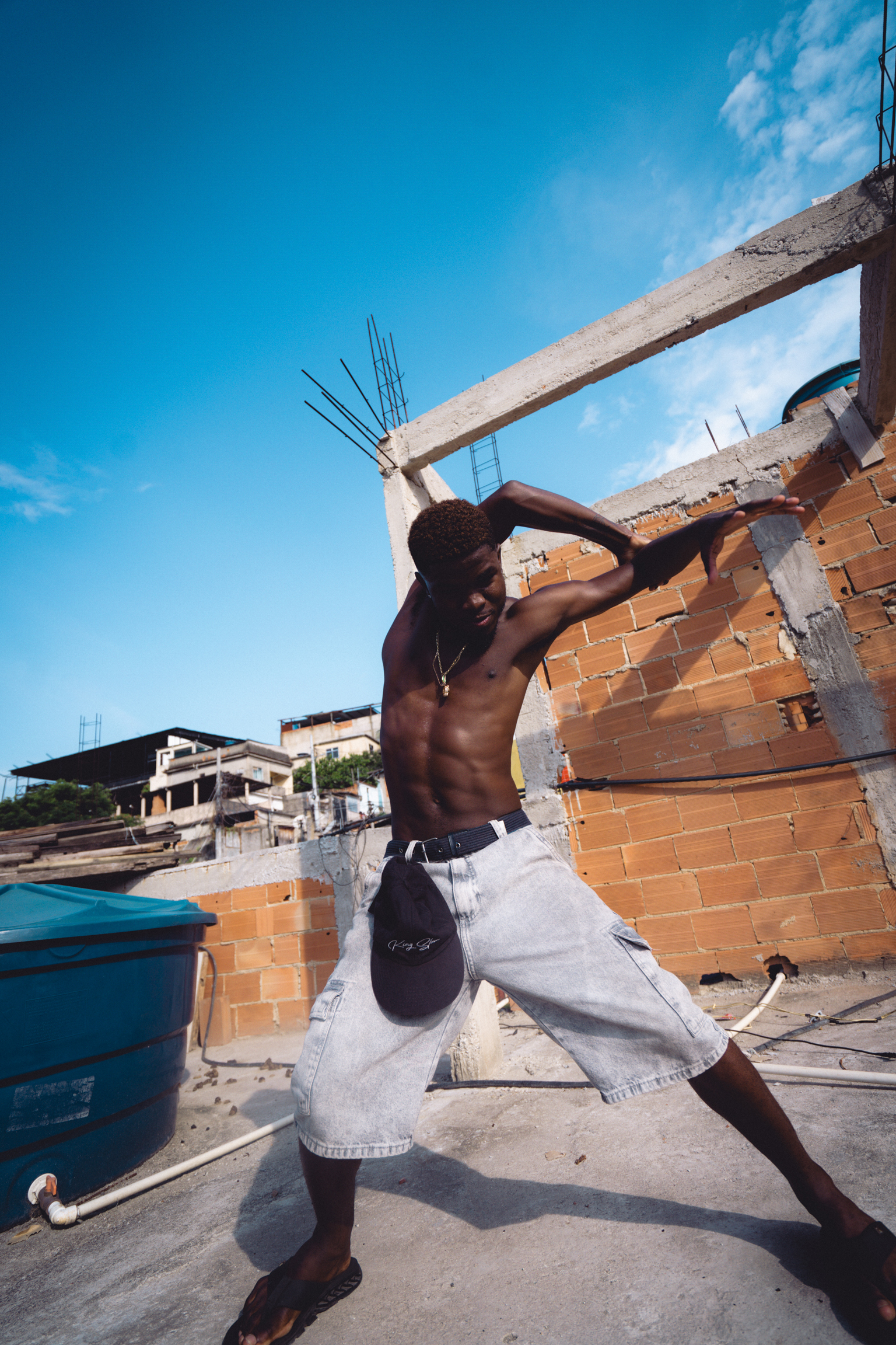 Shirtless man carrying concrete beam at construction site with orange bricks, blue sky, and unfinished buildings in background.