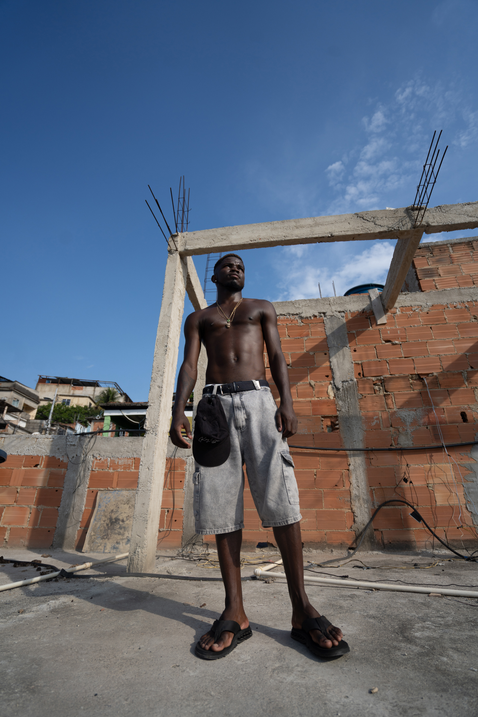 Shirtless man in grey shorts stands on construction site with unfinished brick buildings and concrete pillars under blue sky.