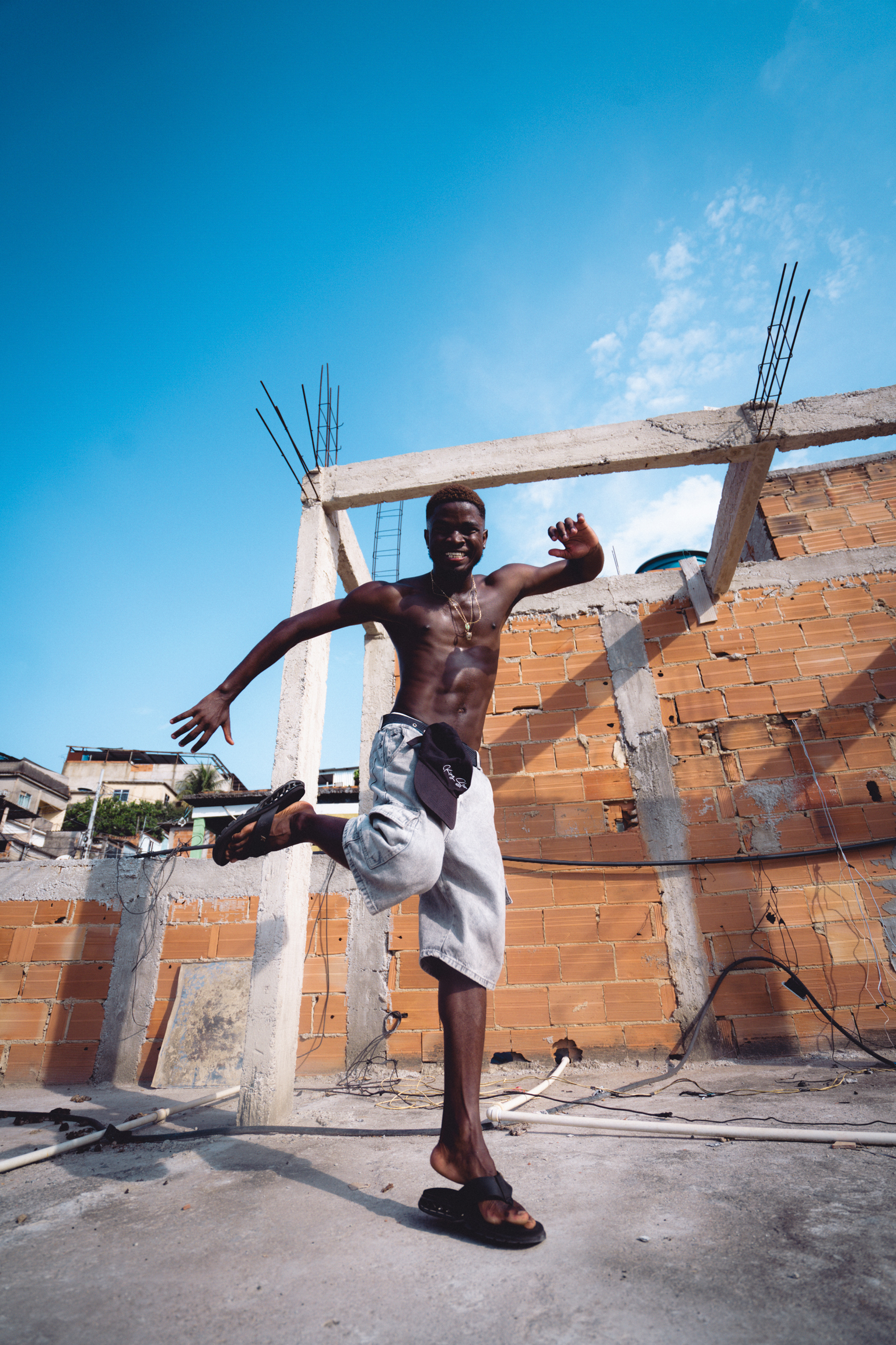 Shirtless man mid-jump at construction site with exposed brick walls, concrete beams and rebar under bright blue sky.