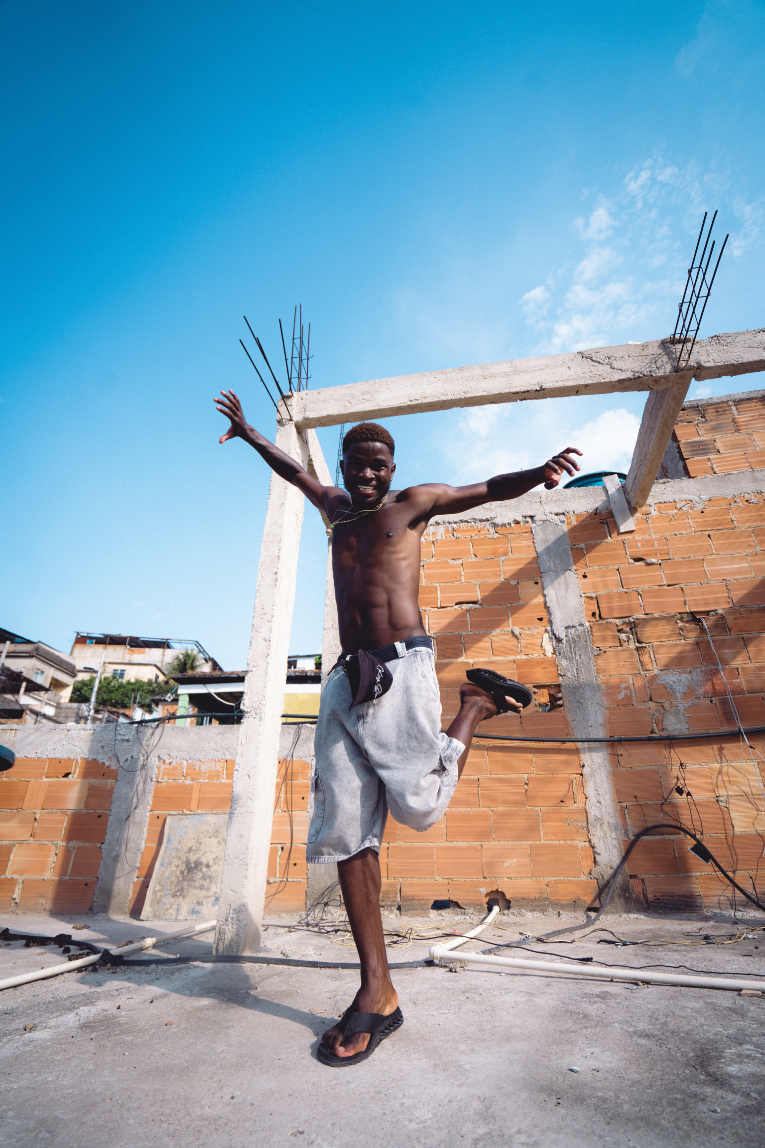 Young man jumping with arms raised at construction site with exposed brick walls and concrete pillars under blue sky.