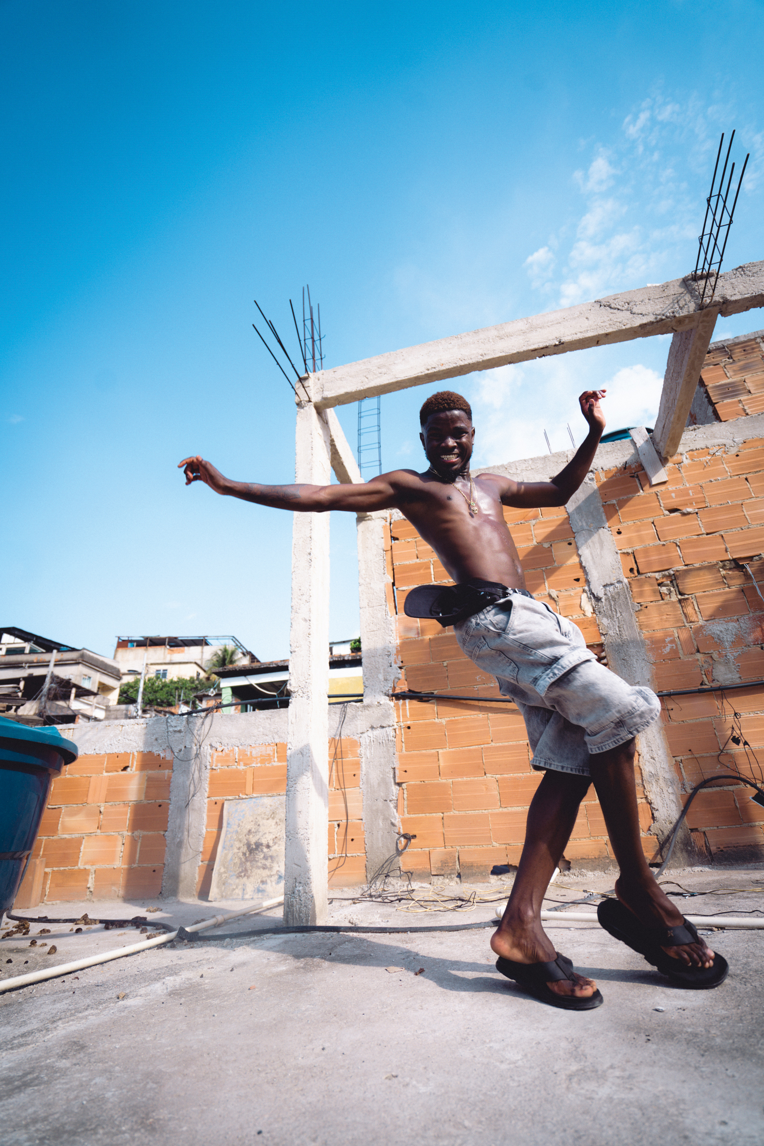 Shirtless man with arms outstretched stands amongst concrete construction beams and orange brick walls under bright blue sky.