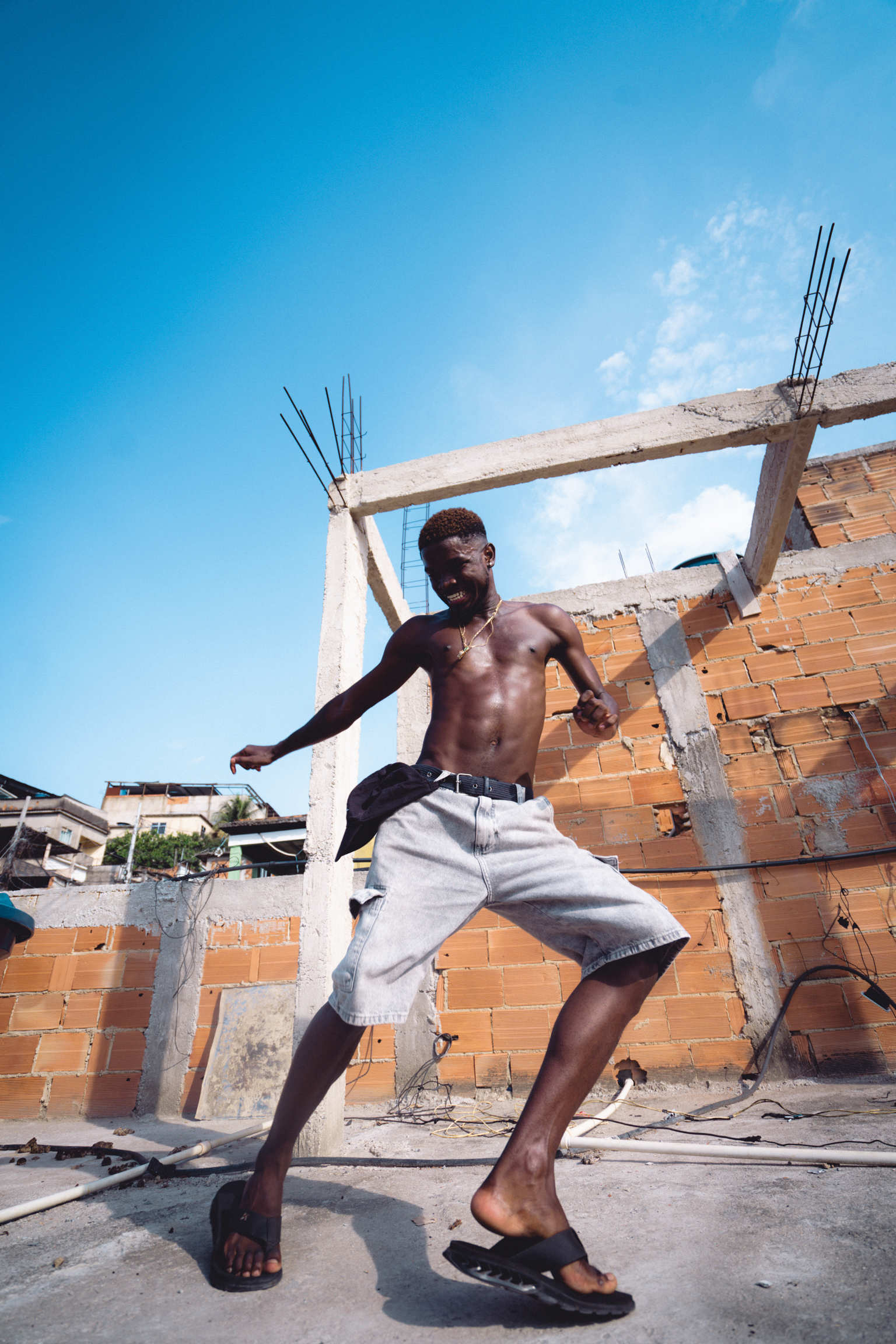 Shirtless man in grey shorts dancing at construction site with exposed brick walls and concrete beams under bright blue sky.