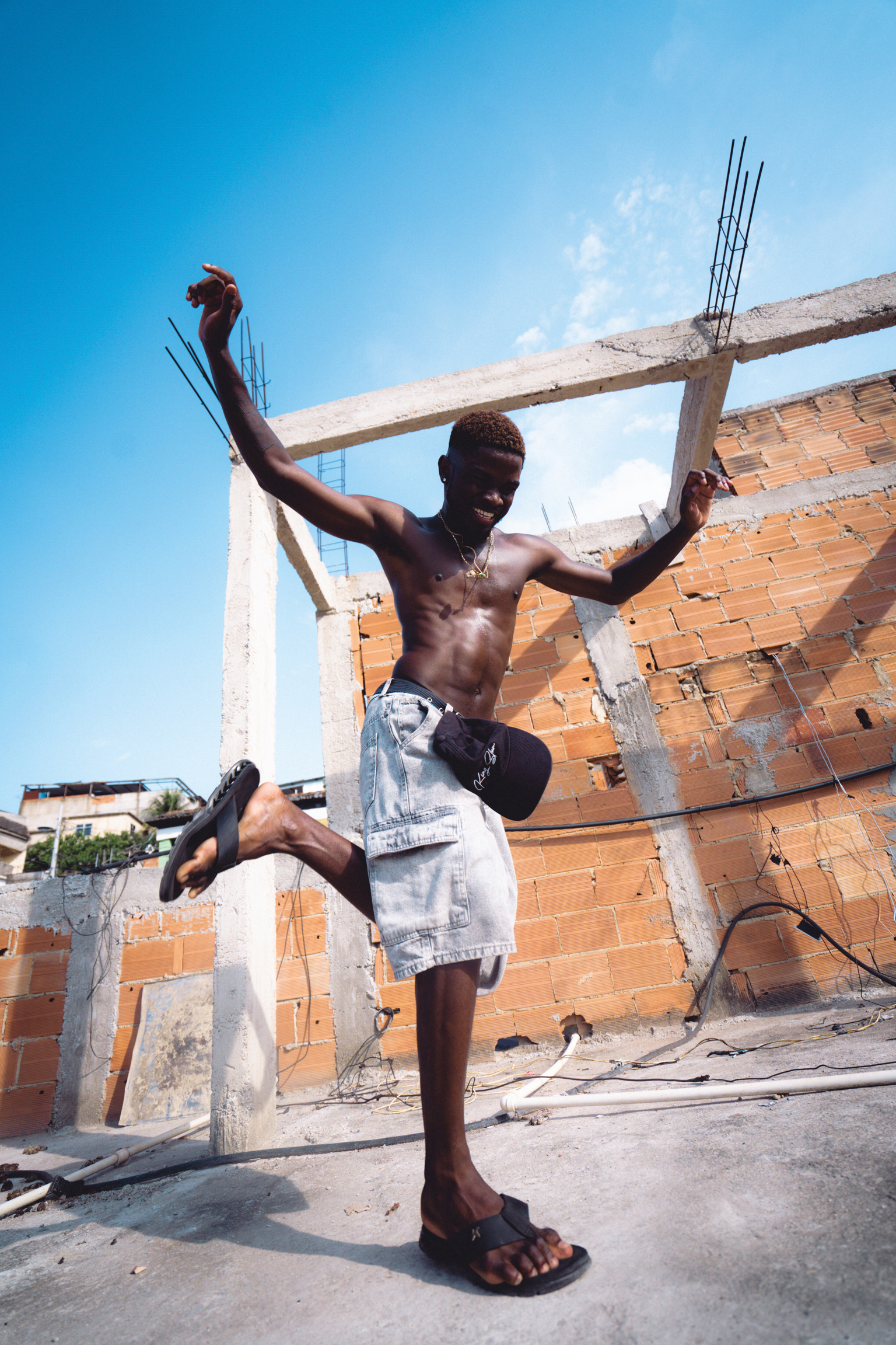 Shirtless man jumping with arms raised at construction site with orange brick walls and concrete beams against blue sky.