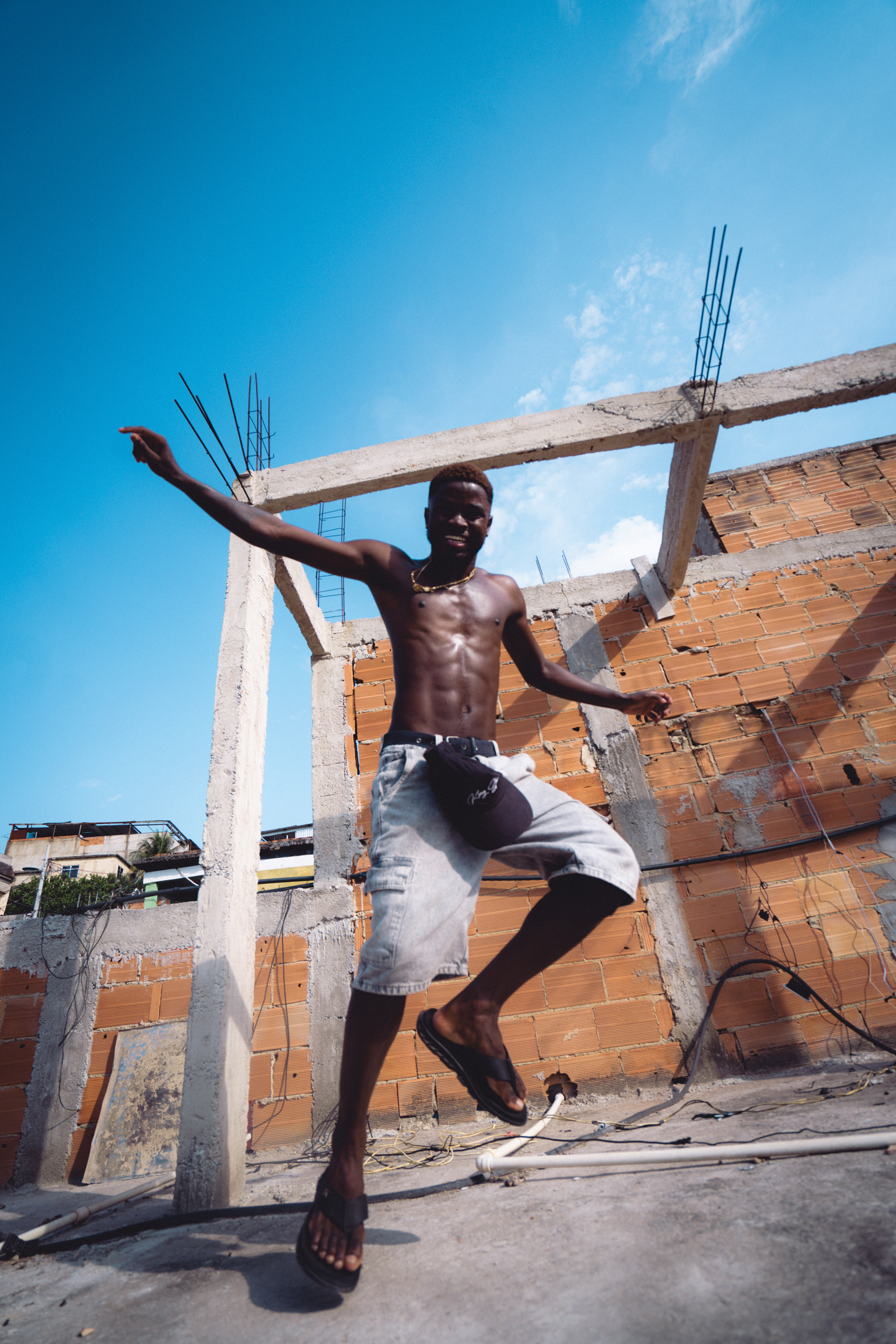 Shirtless man jumping with arms outstretched at construction site with exposed brick walls and concrete beams under blue sky.