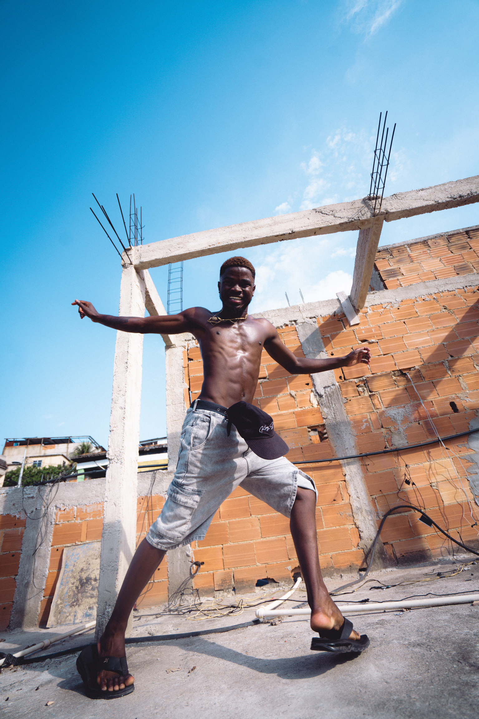 Shirtless man with arms outstretched stands on construction site with exposed brick walls and concrete pillars against blue sky.