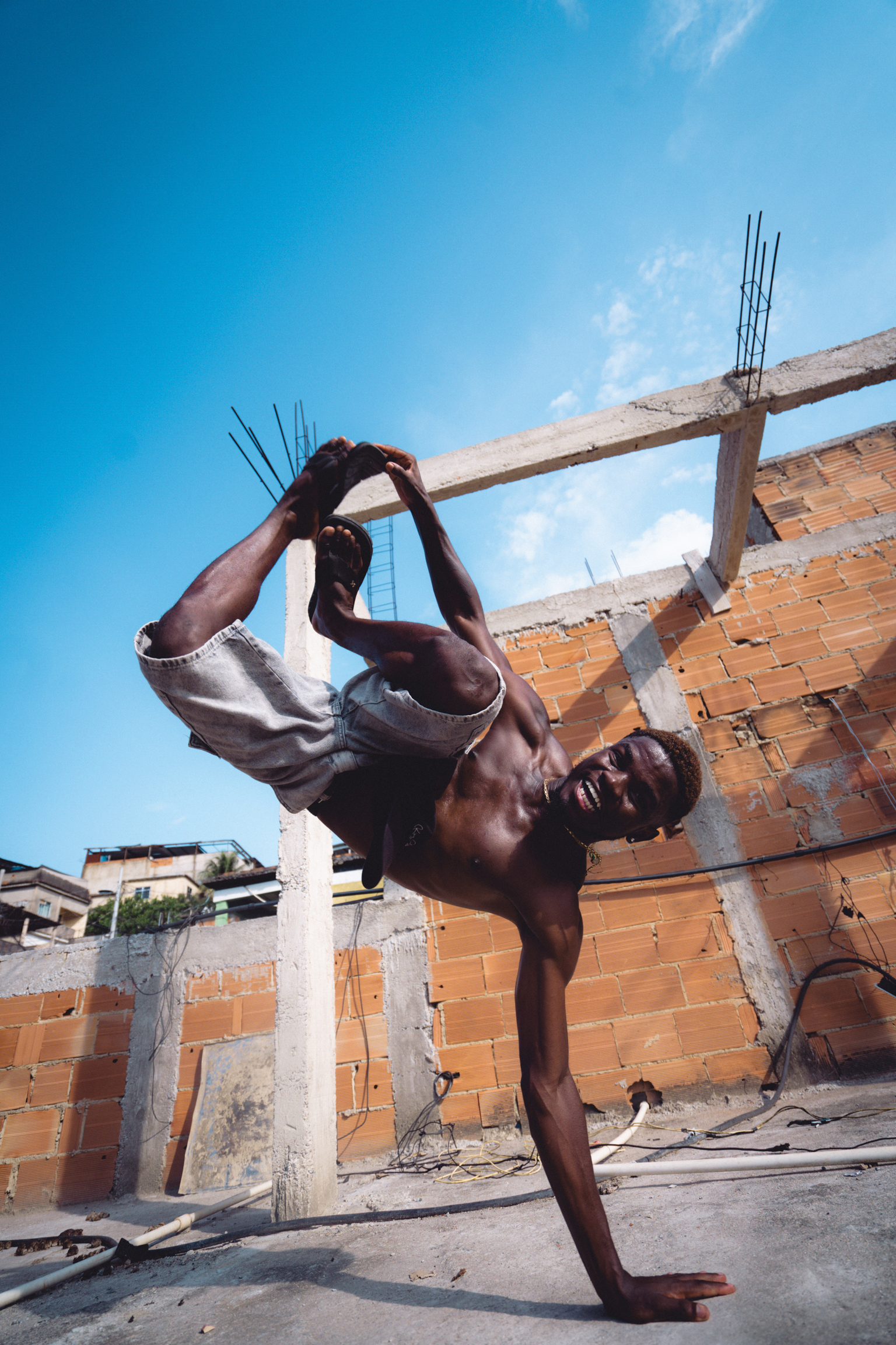 Shirtless man performing handstand on construction site with exposed brick walls and concrete beams against blue sky.