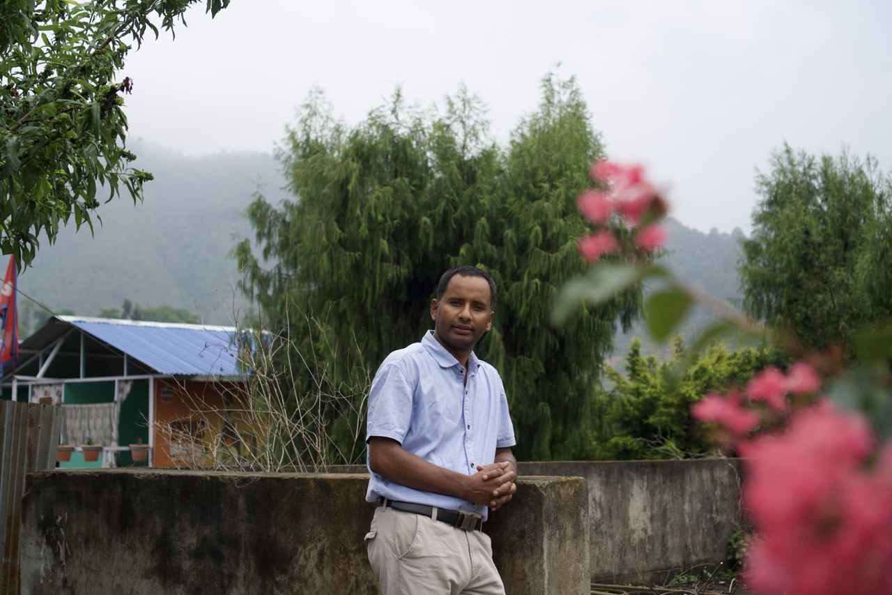 Man in white shirt standing by concrete wall with pink flowers in foreground, green trees and hills in background.