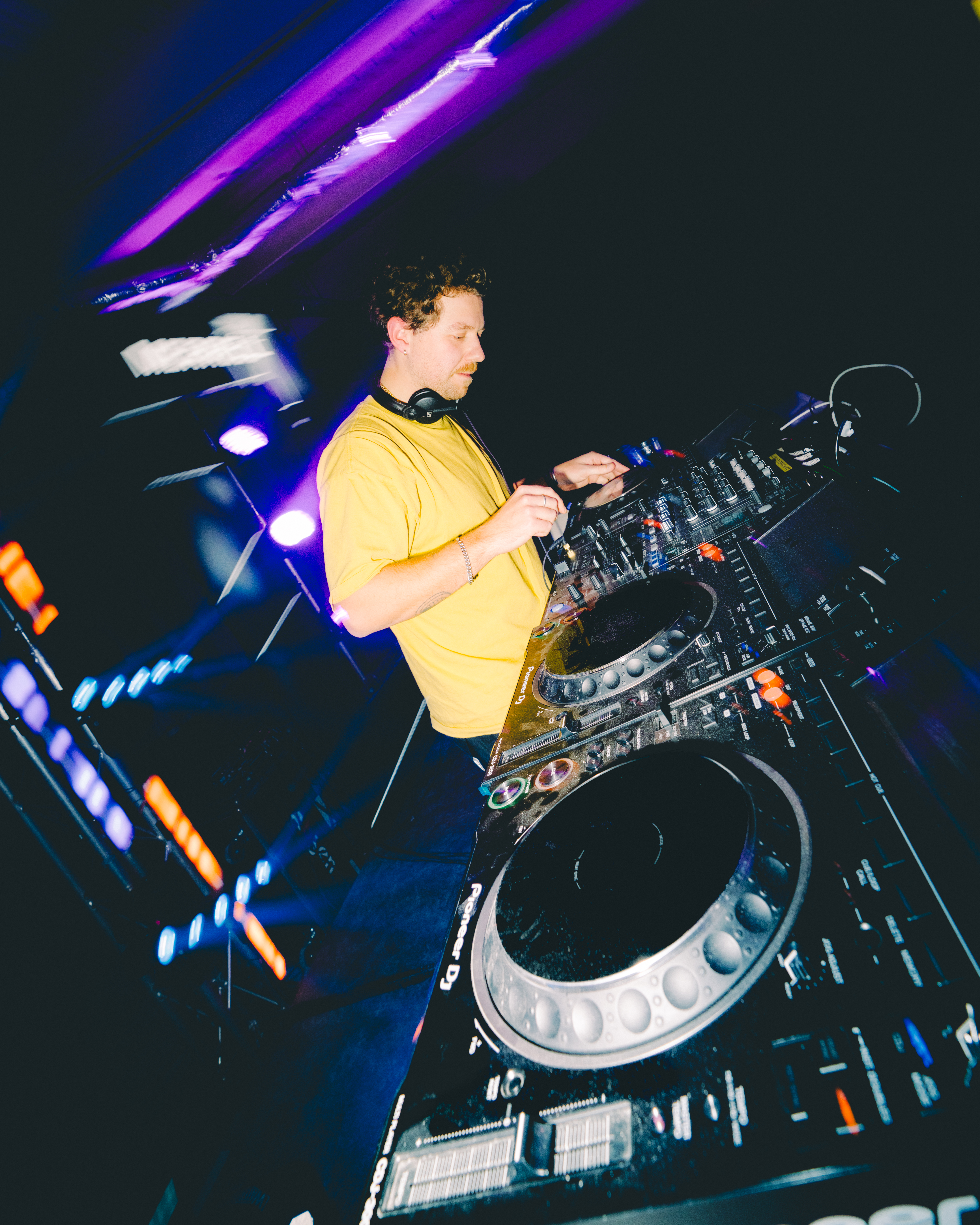 DJ in yellow shirt operating mixing deck and turntables under purple and blue stage lighting with motion blur effects.