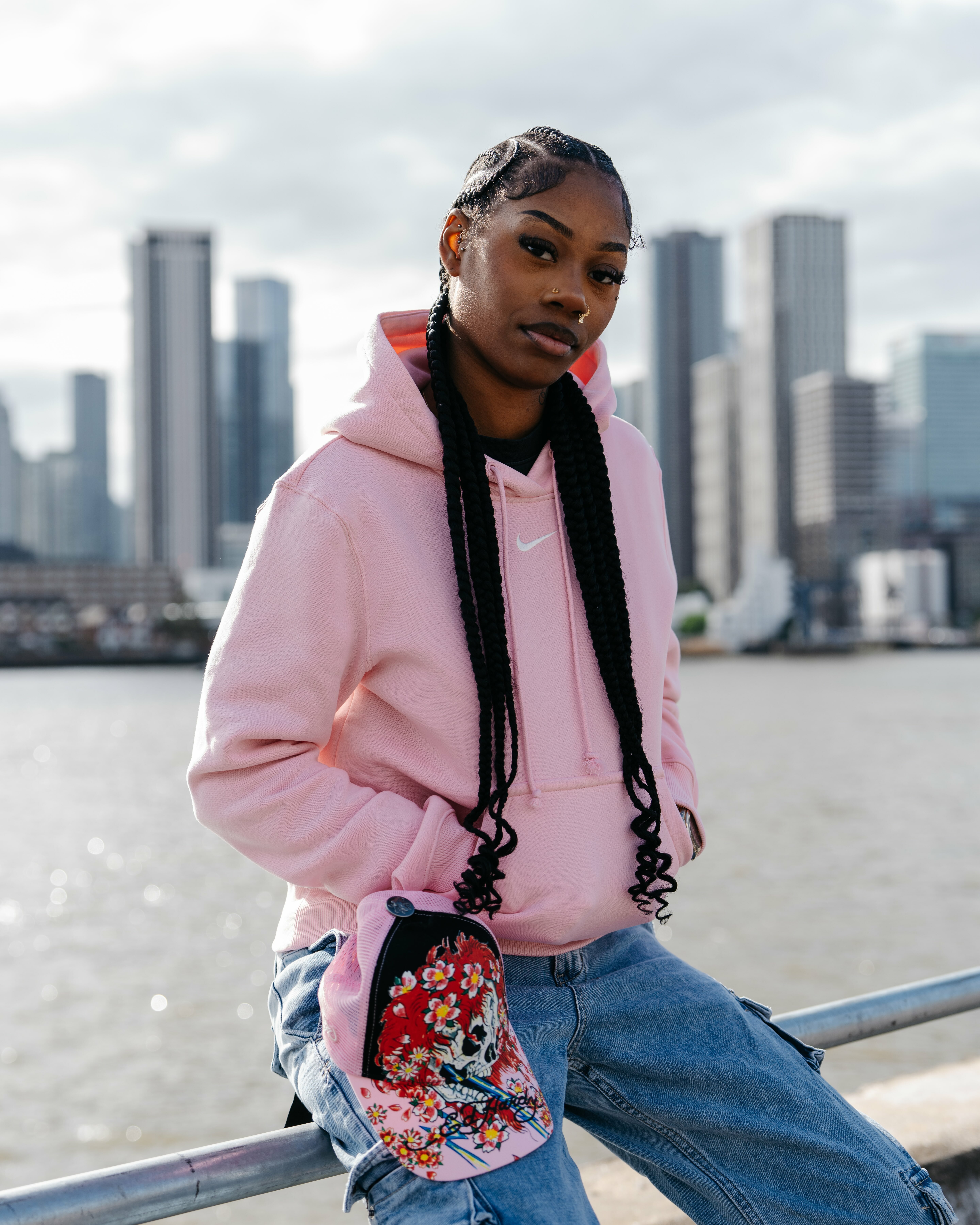 Woman with long braids wearing pink hoodie and jeans, sitting by waterfront with city skyline in background.