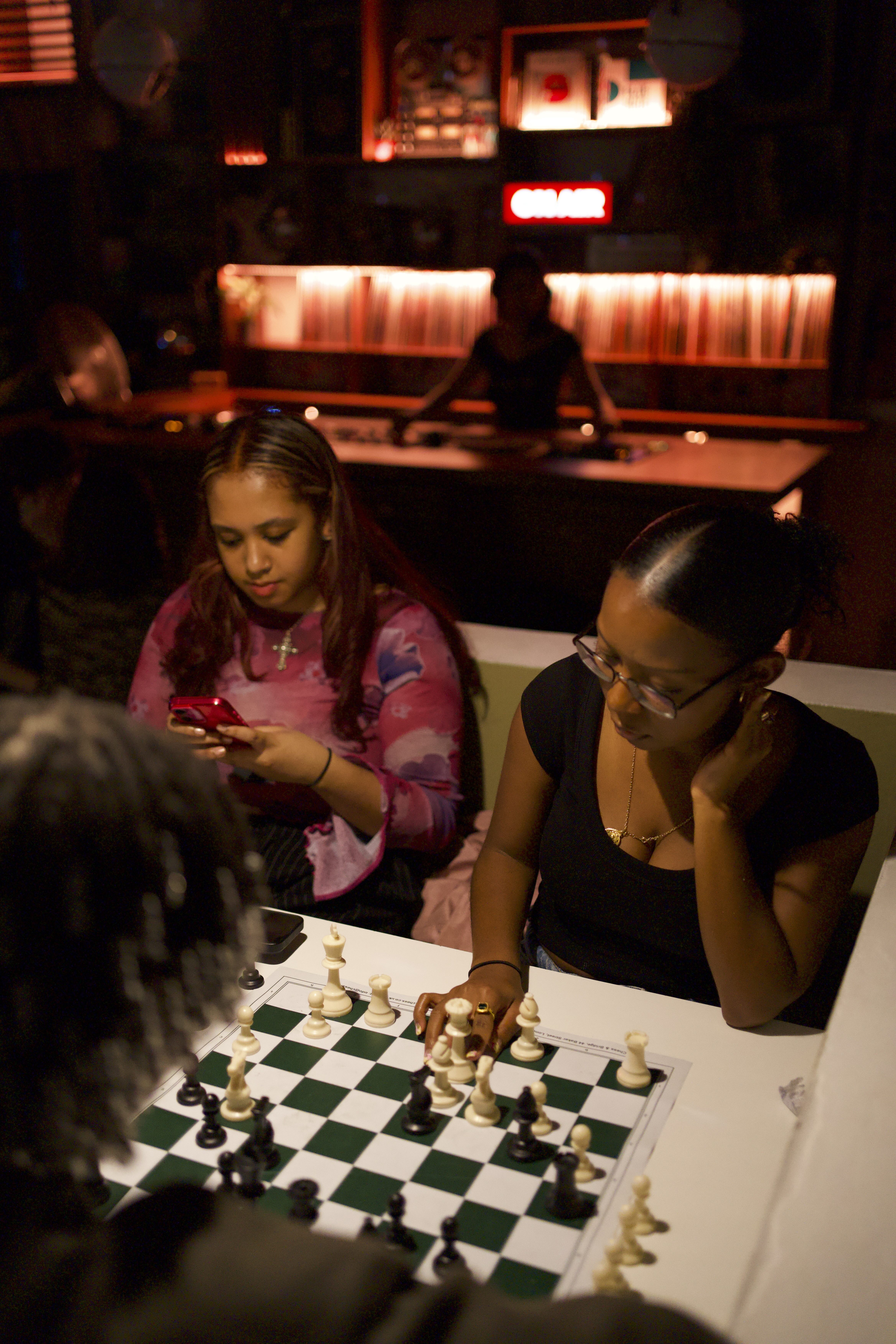 Two people at white table with green and white chessboard between them in dimly lit bar with red neon lighting in background.