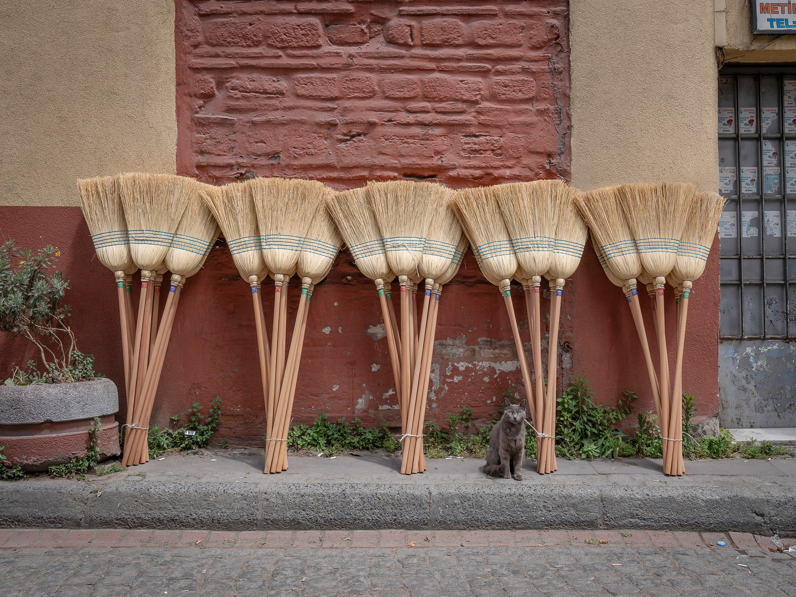 Five straw brooms with wooden handles leaning against red brick wall on pavement outside building with metal-grilled window.