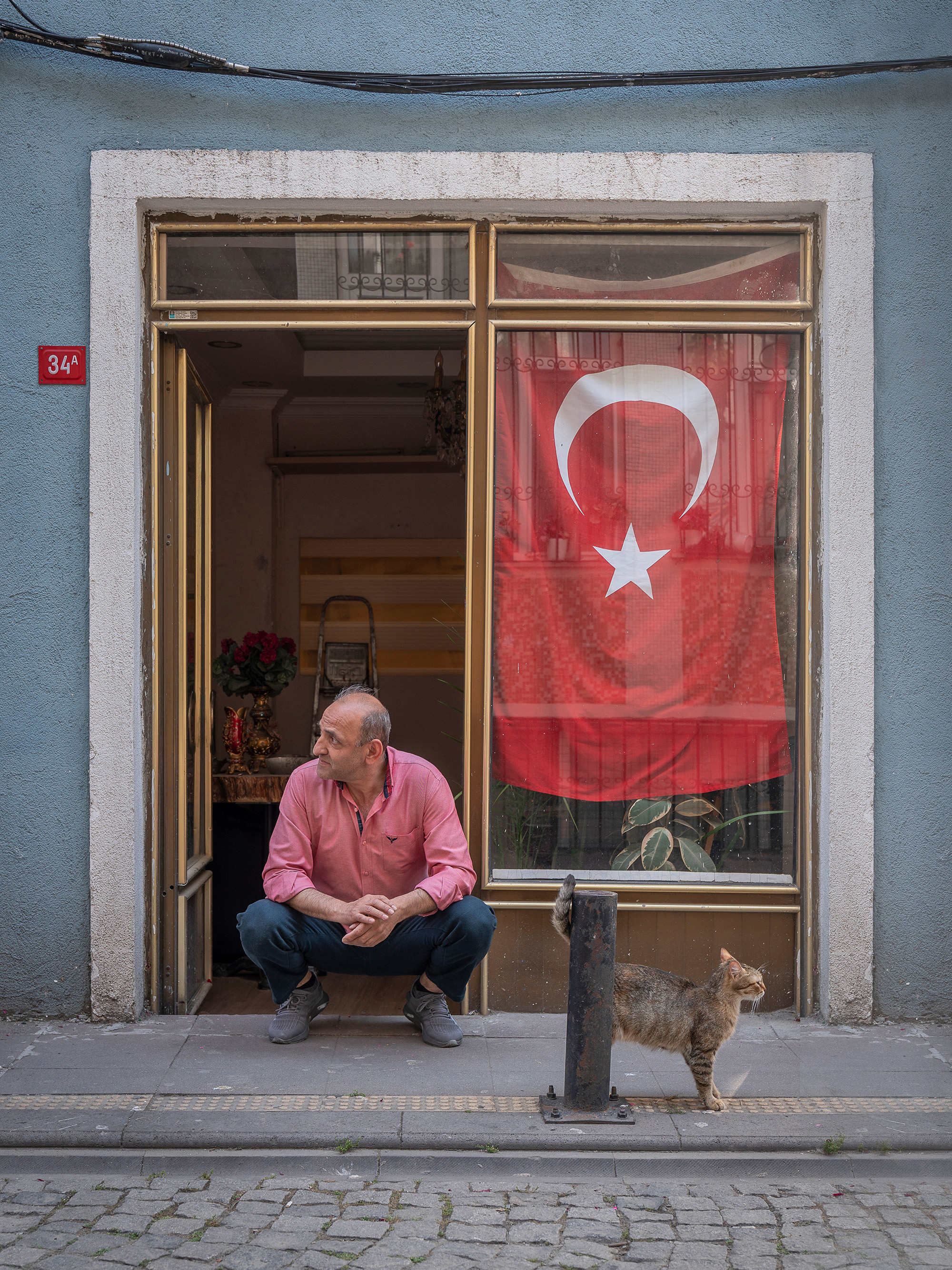 Elderly man in pink shirt sits in doorway of blue building with Turkish flag displayed in window, tabby cat stands nearby on pavement.