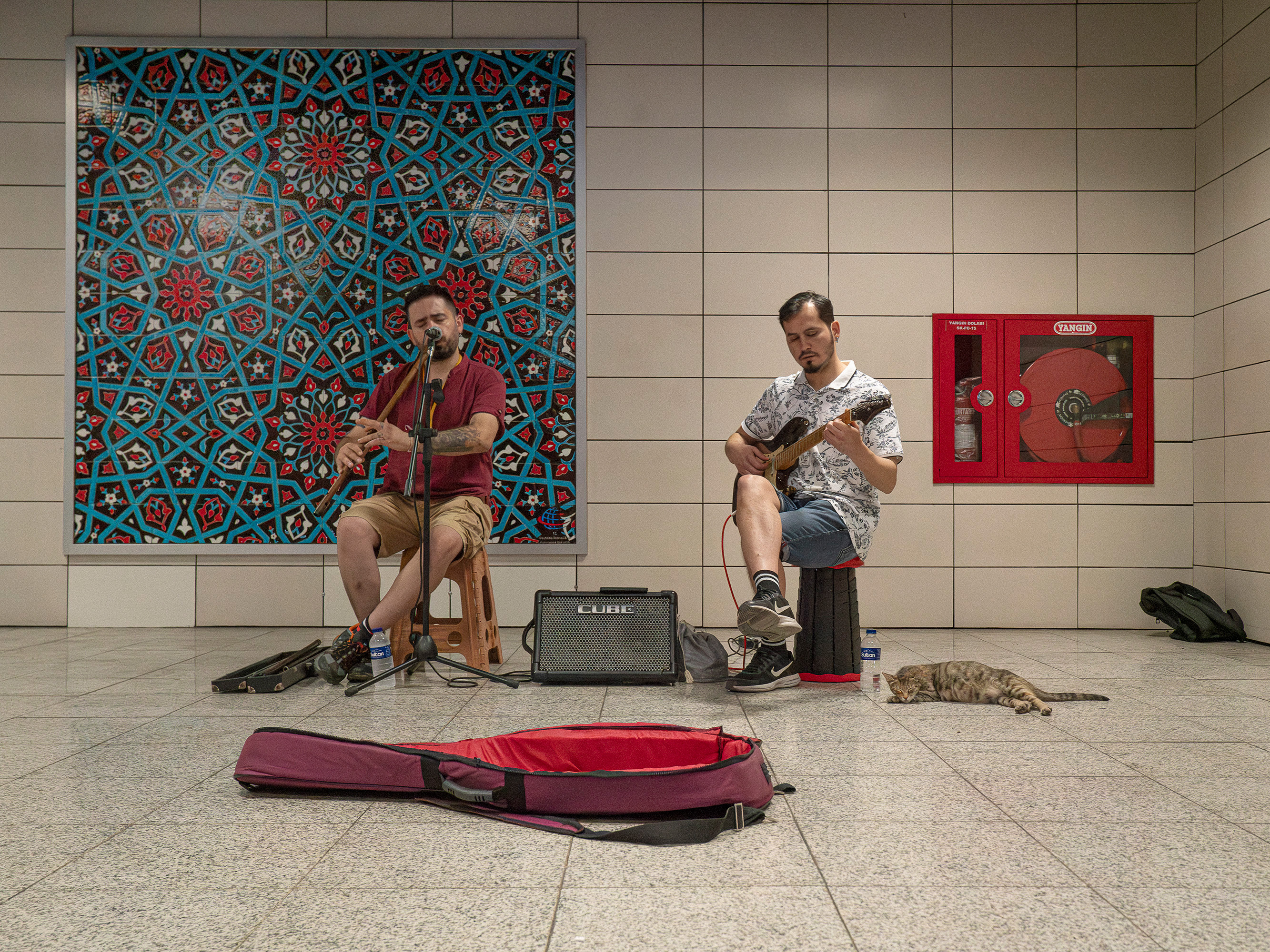 Two men busking in underground station - one plays flute, other holds guitar. Colourful geometric mosaic behind them, red guitar case open.