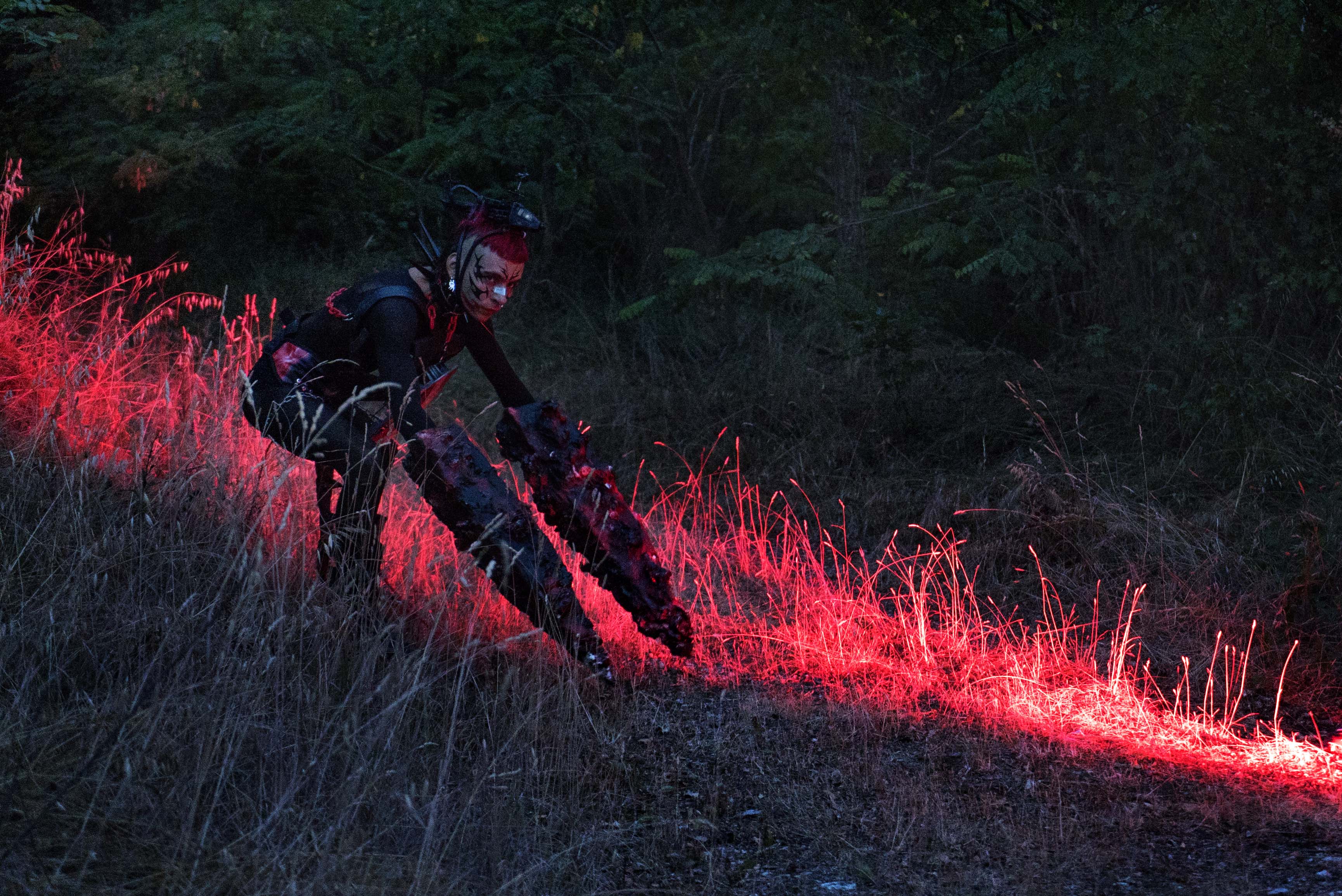 Person in dark clothing using angle grinder creating bright red sparks against dark background with trees visible above.