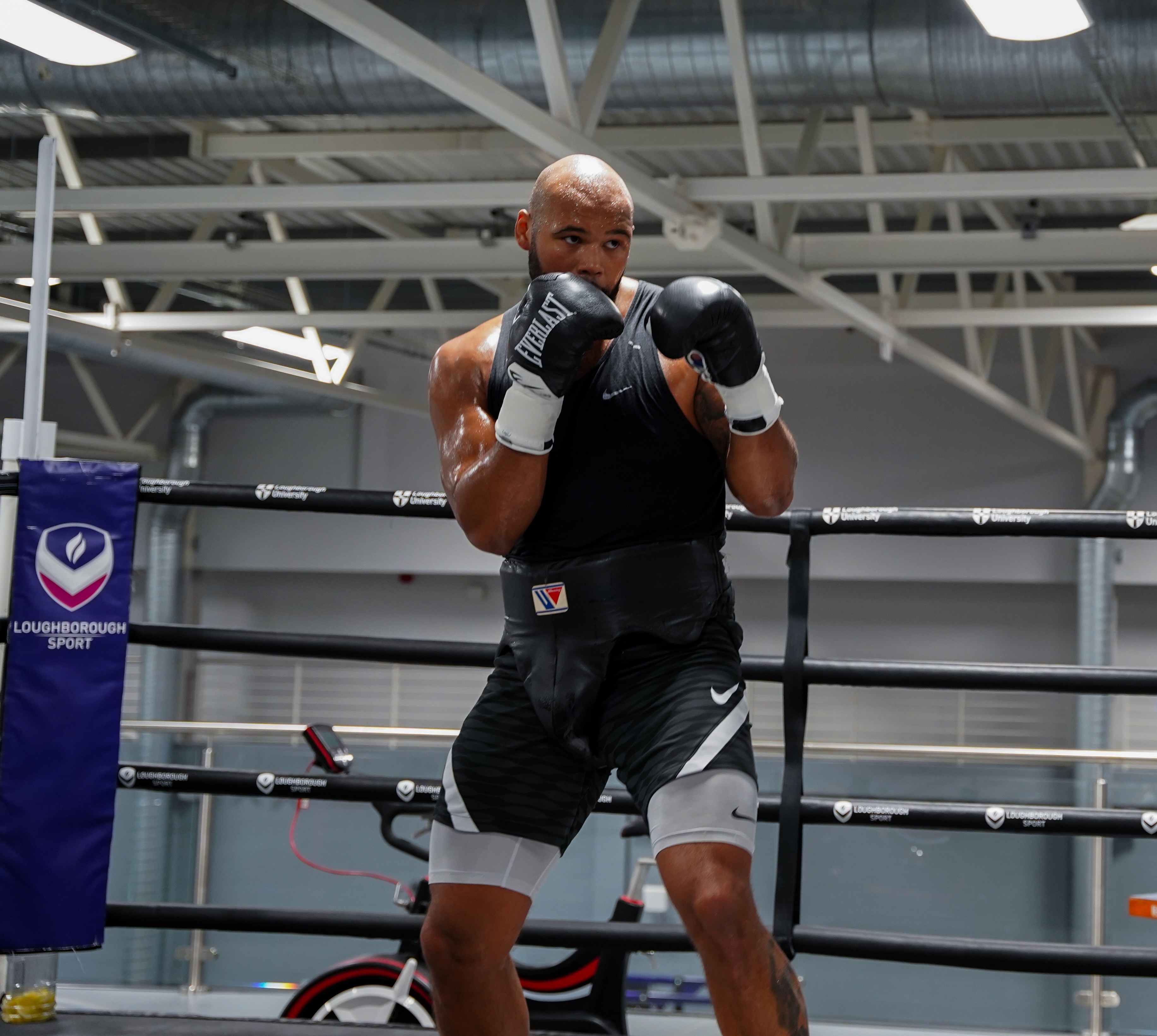 A muscular man wearing boxing gloves and a black outfit, standing in a boxing ring surrounded by equipment.