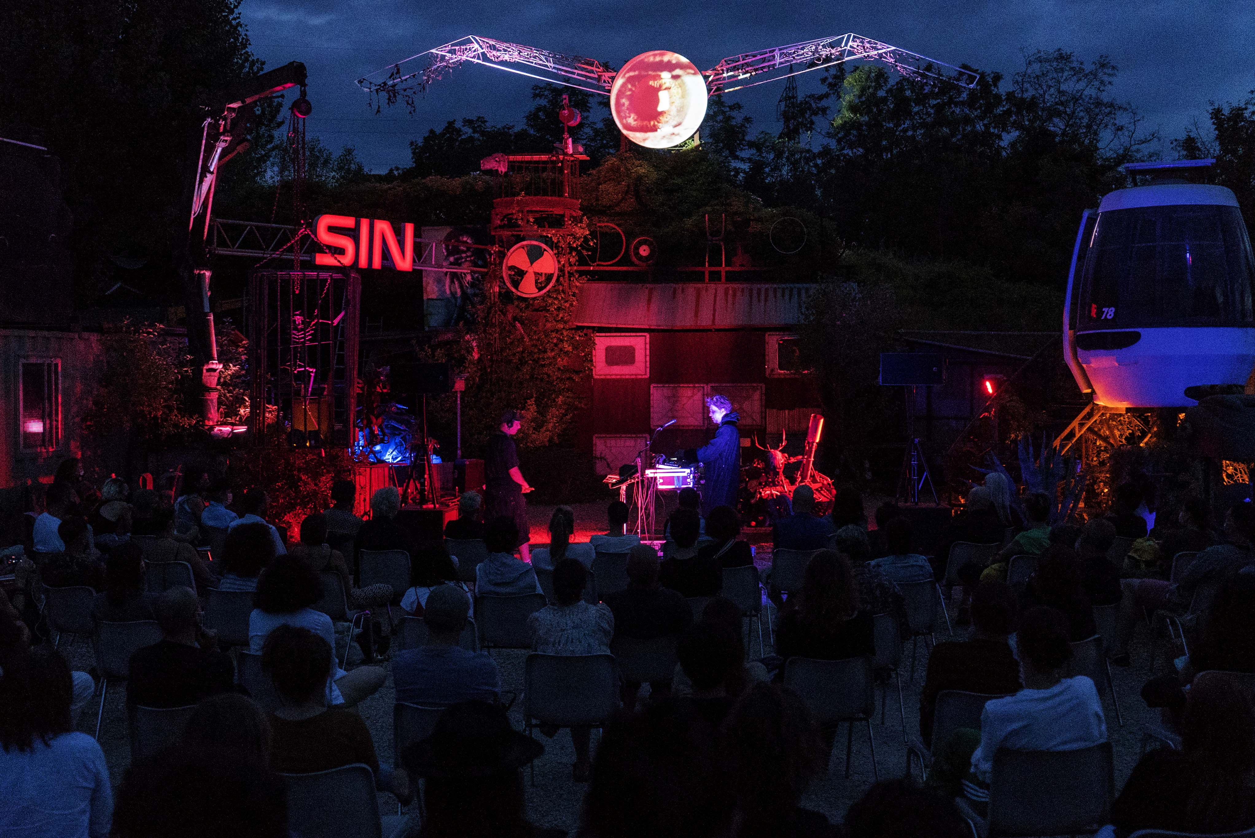 Outdoor venue at night with red neon lighting, crowd gathered below illuminated stage area with "SIN" sign visible against dark blue sky.