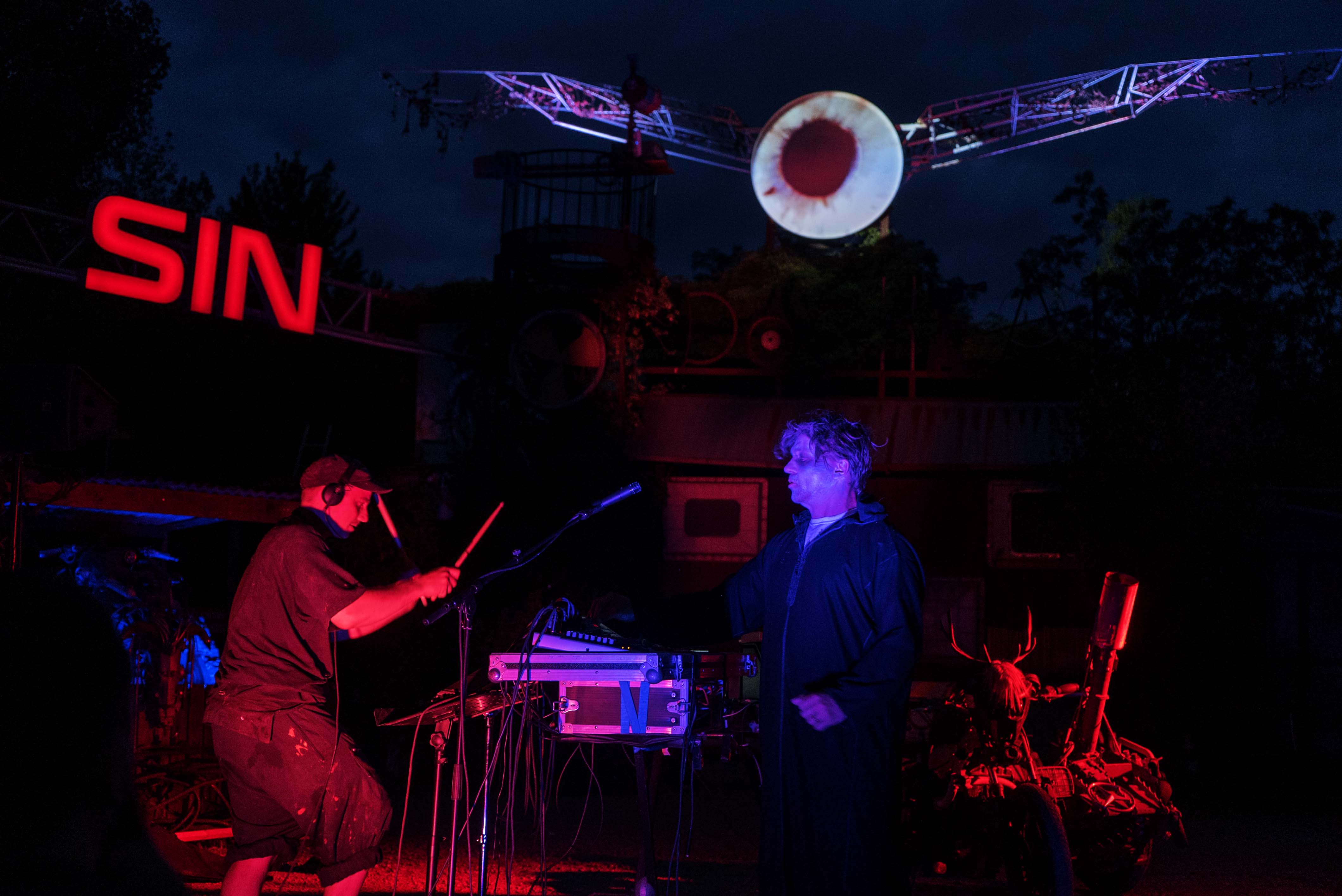 Dark stage with red and blue lighting. Musicians performing with keyboards and equipment. Red "SIN" sign and white circular projection above.