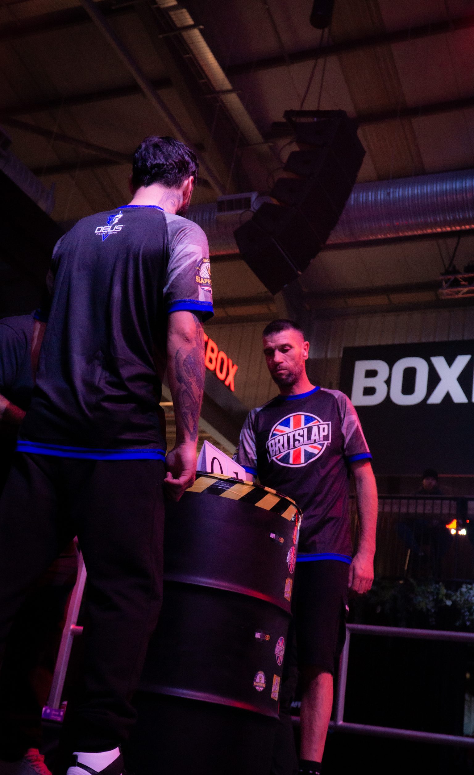 Two men in boxing gym, one standing on elevated platform, other below wearing Union Jack shirt, industrial ceiling with exposed beams.
