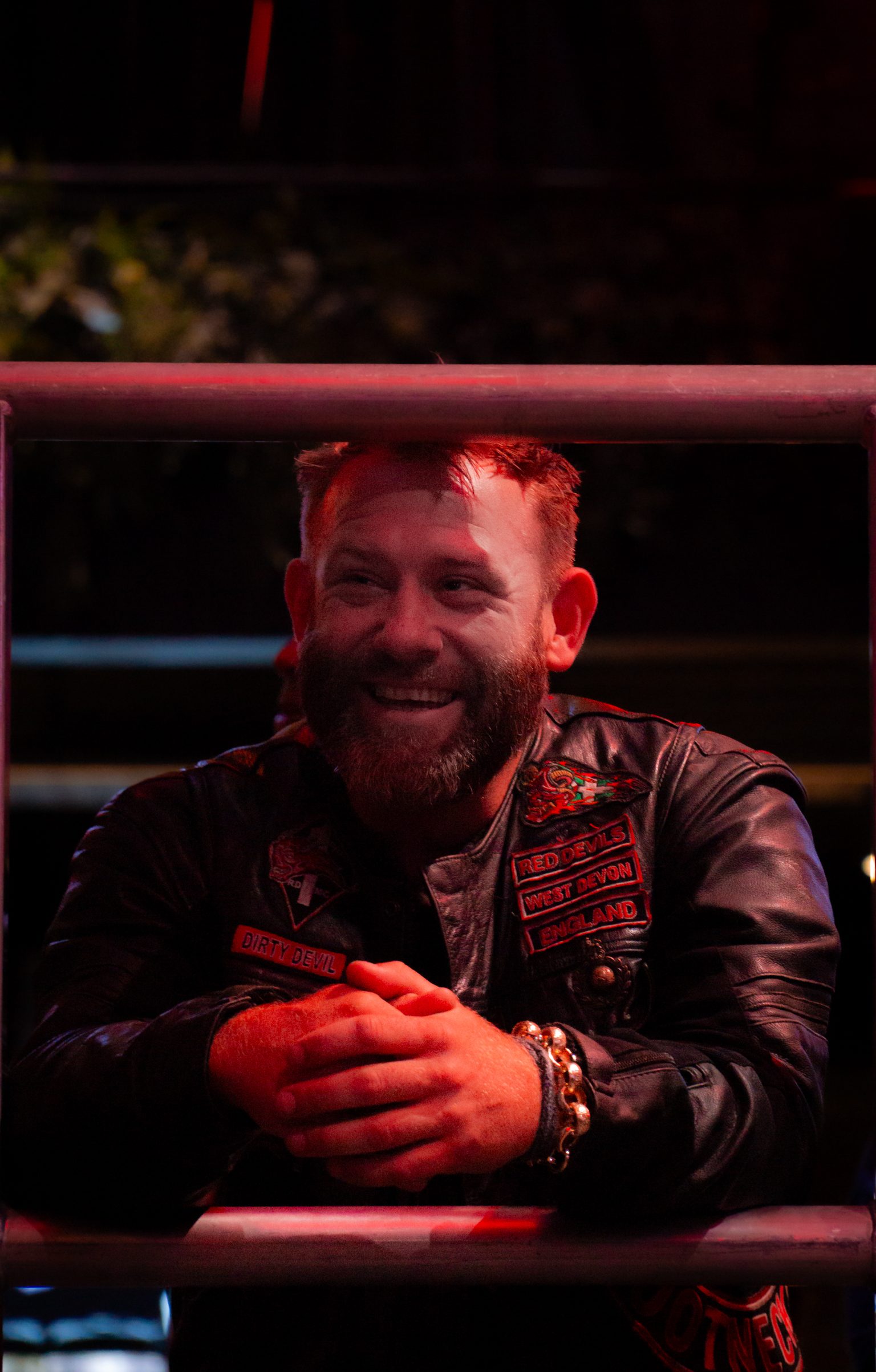 Bearded man in leather jacket with patches smiling, leaning on rope barrier under red lighting in dark venue.