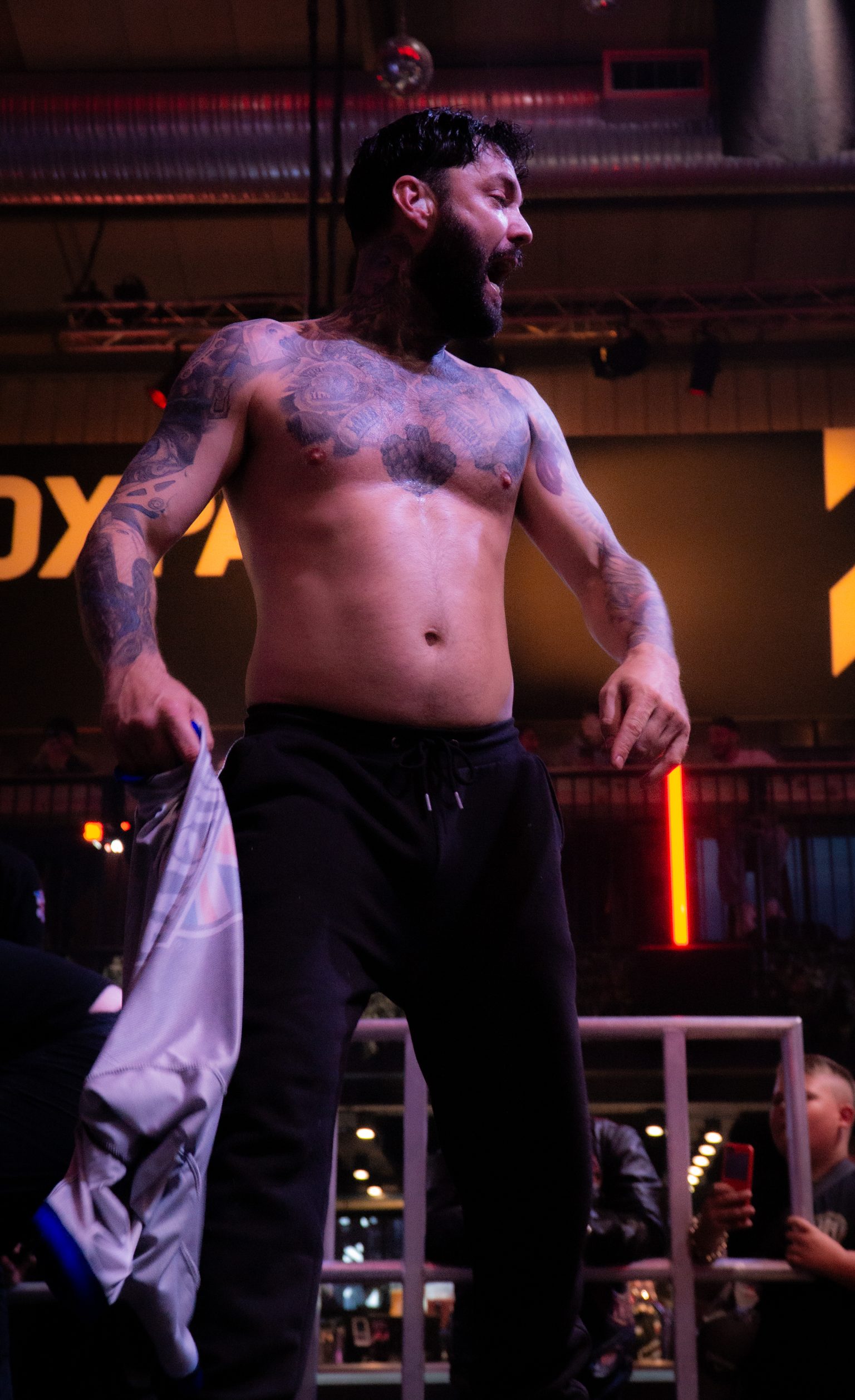 Shirtless tattooed wrestler with dark beard holding white garment, standing on platform with orange lighting and crowd below.