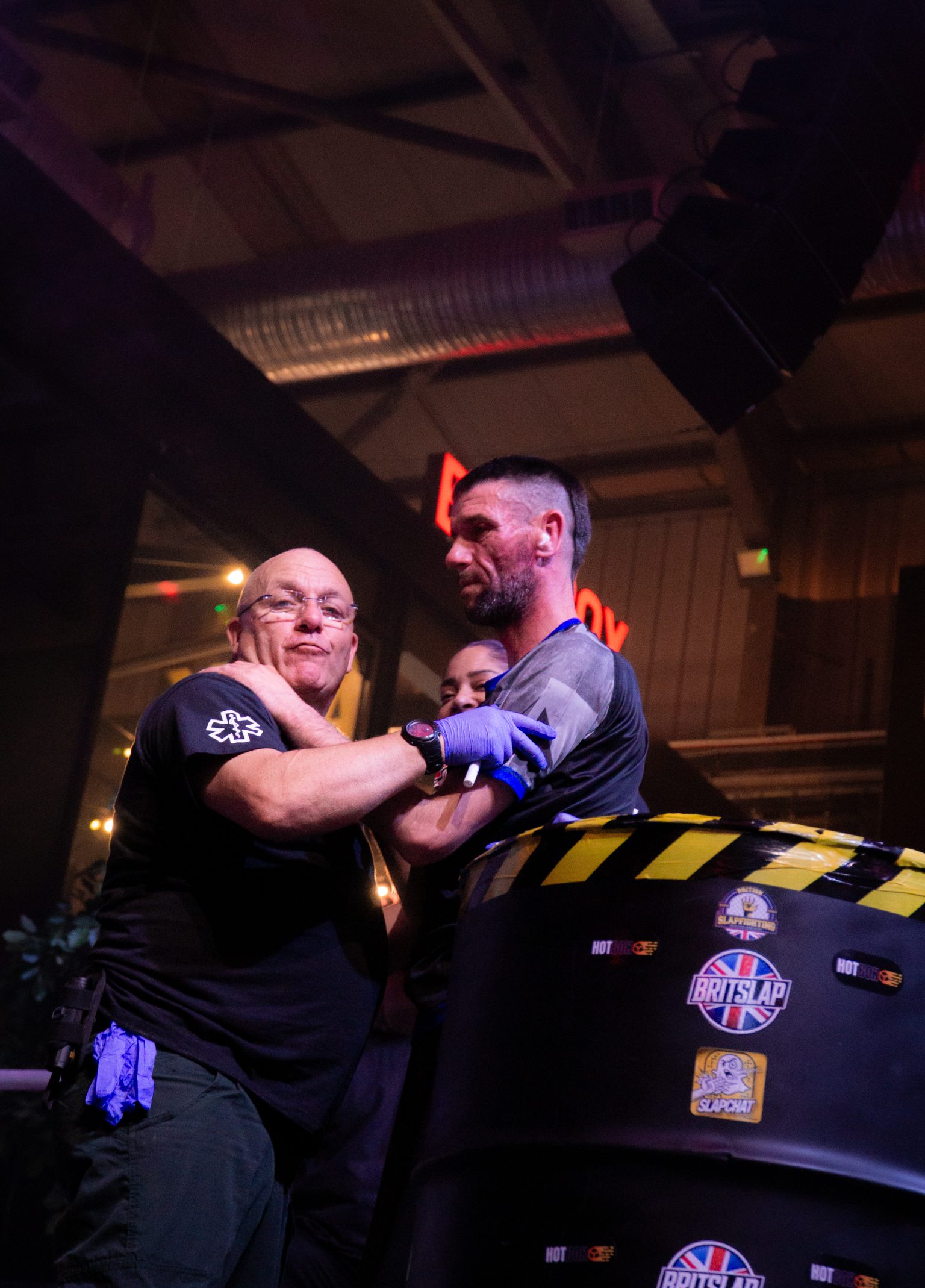 Two men arm wrestling at table with yellow and black striped edge in dimly lit venue with industrial ceiling and warm lighting.