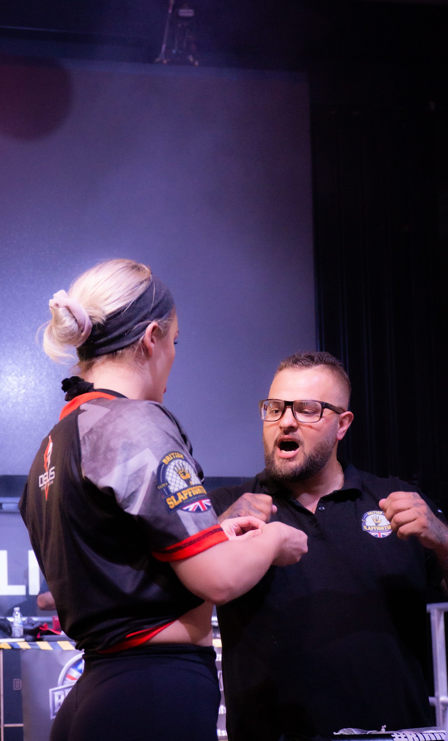 Bearded man in glasses and black polo shirt speaking with blonde woman in grey sports shirt with red trim on stage.