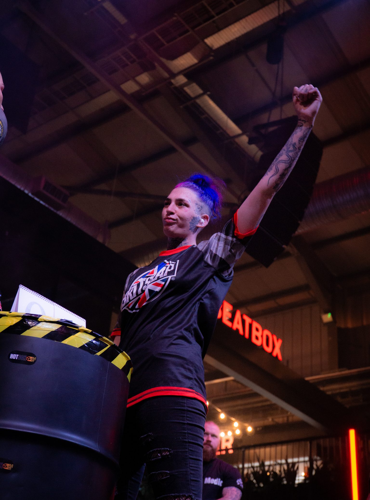 Woman in black jersey raises fist triumphantly at indoor venue with exposed ceiling and red "BEATBOX" sign in background.