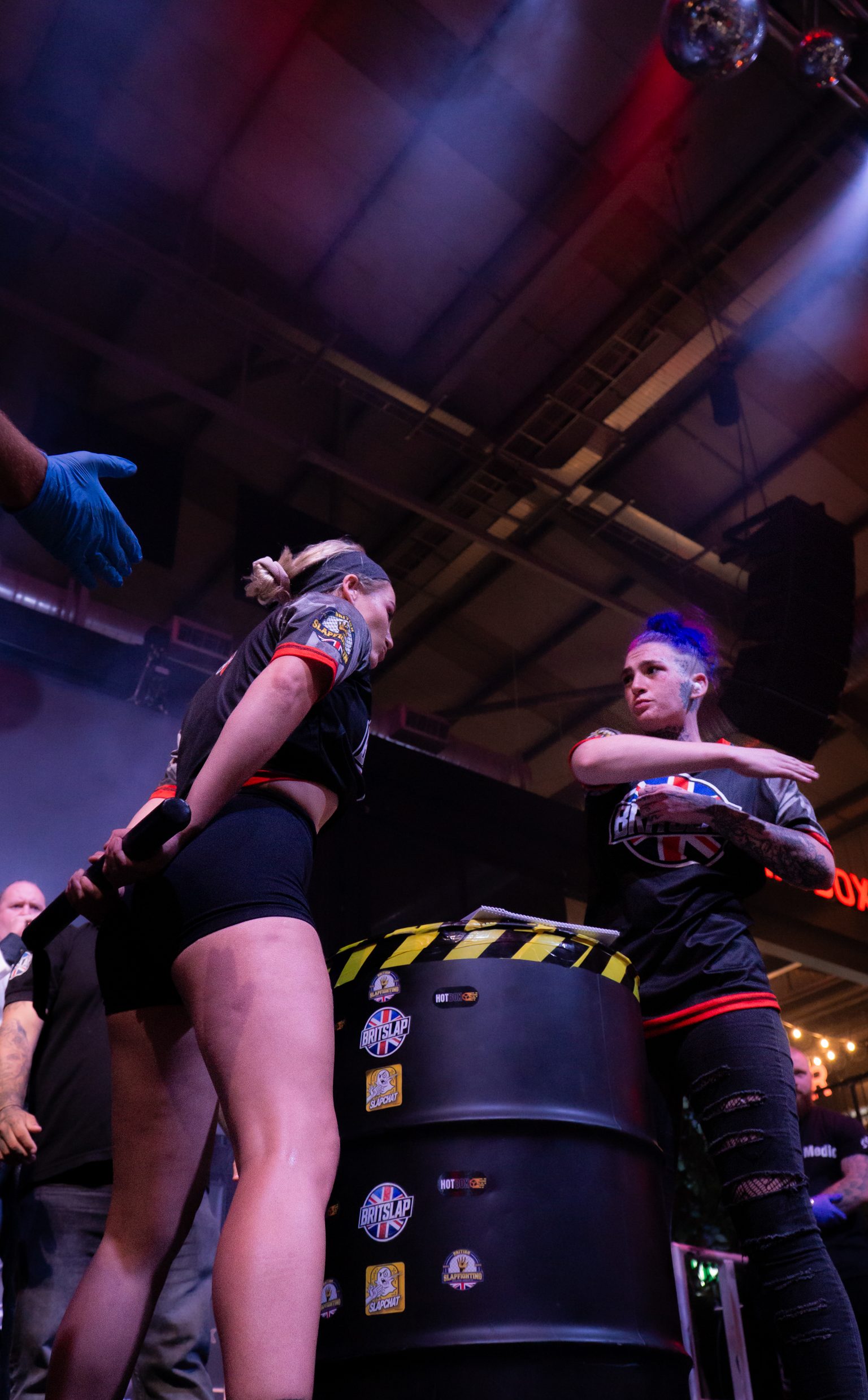 Two people in dark clothing at industrial venue with exposed ceiling beams, blue and red stage lighting, disco ball visible above.