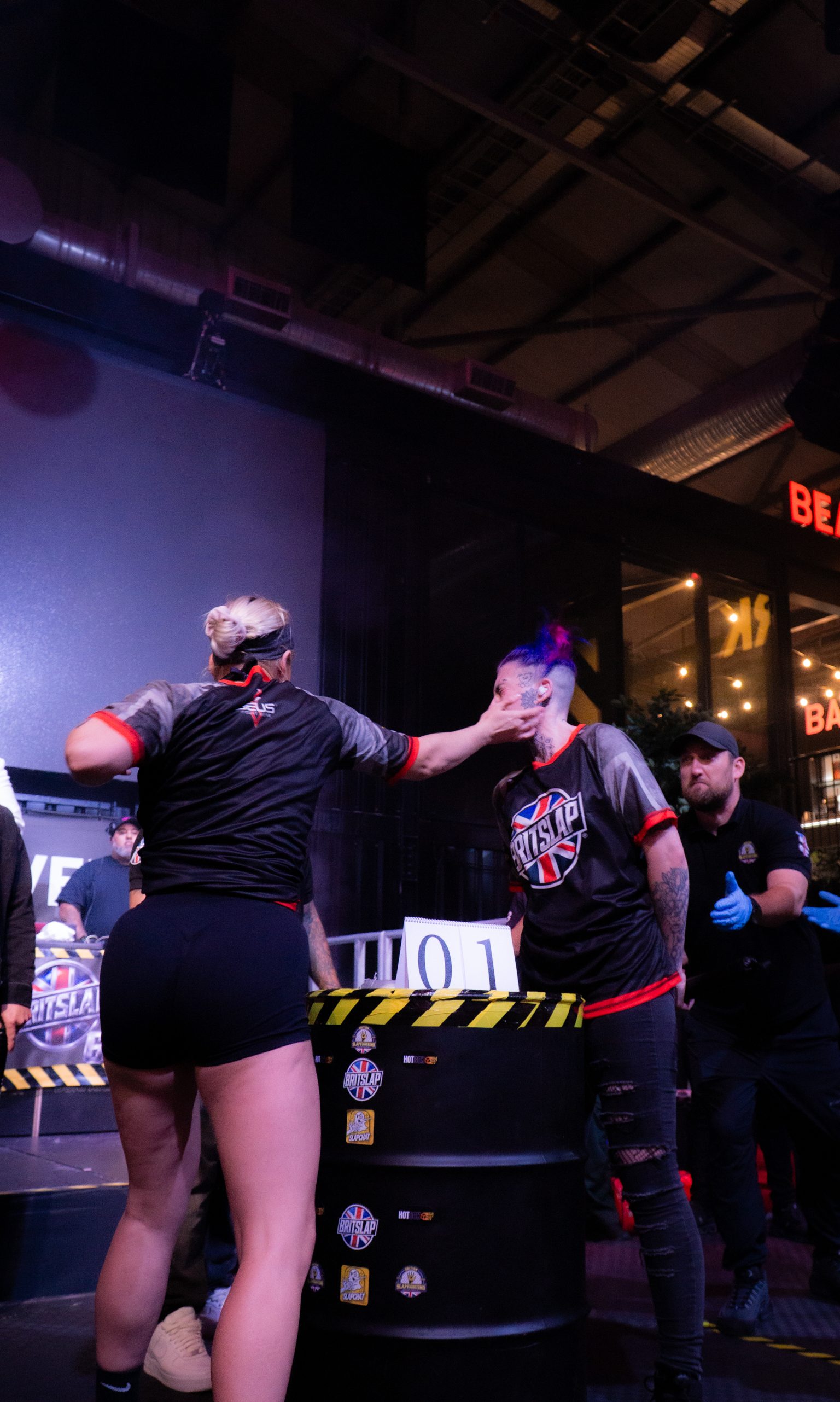 Two slap fighters competing at black table with yellow striped barrier, referee in background, purple lighting overhead.