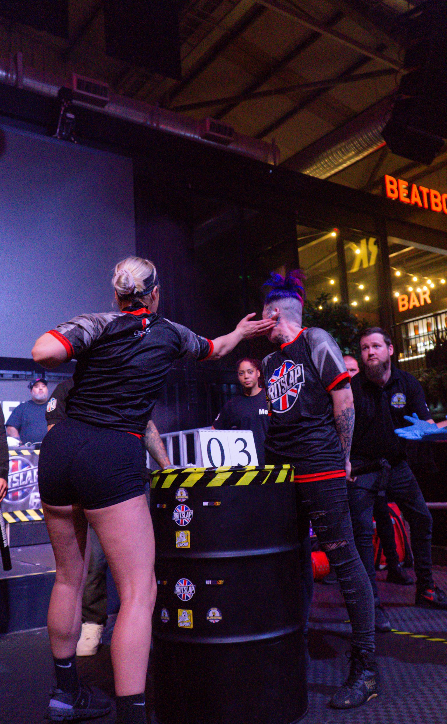 Two women in black sports kit stand beside black equipment marked "03" in industrial venue with purple lighting and exposed ceiling beams.