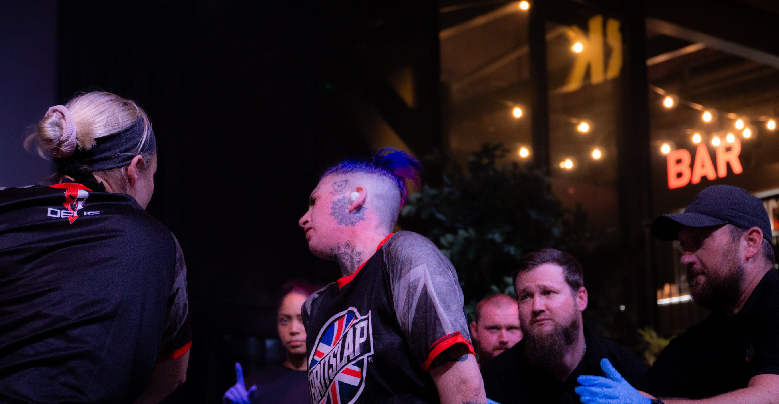 Police officers arresting tattooed man with pink hair outside illuminated bar at night, crowd gathered around.