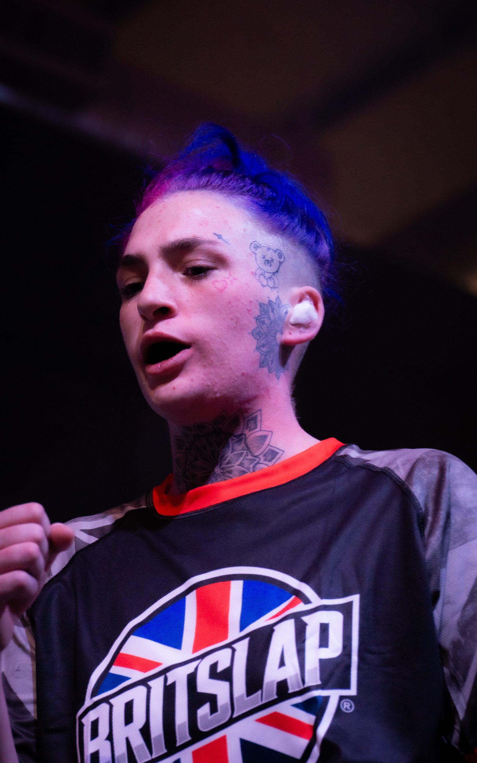 Woman with blue hair and neck tattoos wearing black Britslap shirt with Union Jack logo under purple and blue stage lighting.